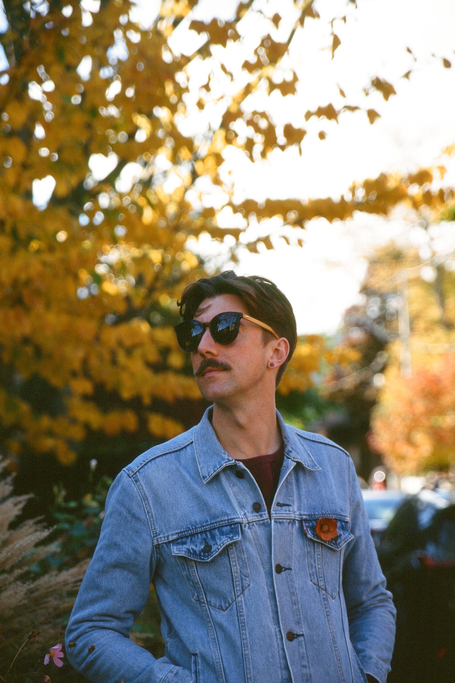 A man poses for a portrait photo wearing sunglasses and a denim shirt. He is combing his moustache in front of a tree with yellow leaves.
