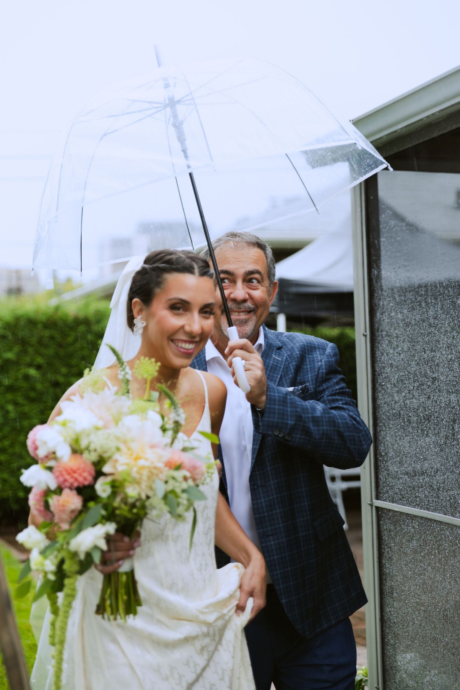 Bride and father of the bride walking to the ceremony together with a clear umbrella held overtop of them.