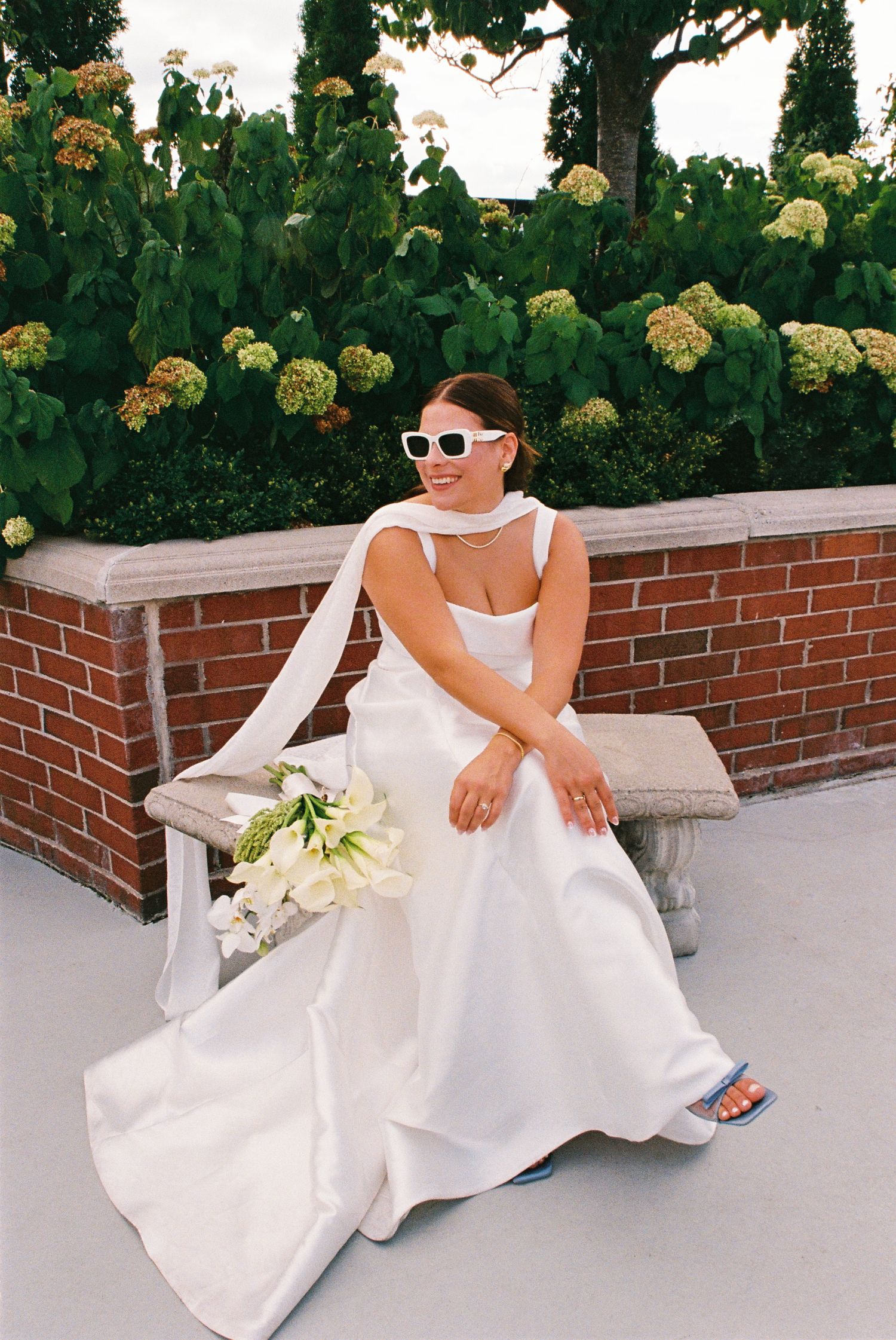 Bride sitting in the rooftop garden wearing an all white satin dress and scarf and retro white and black sunglasses.