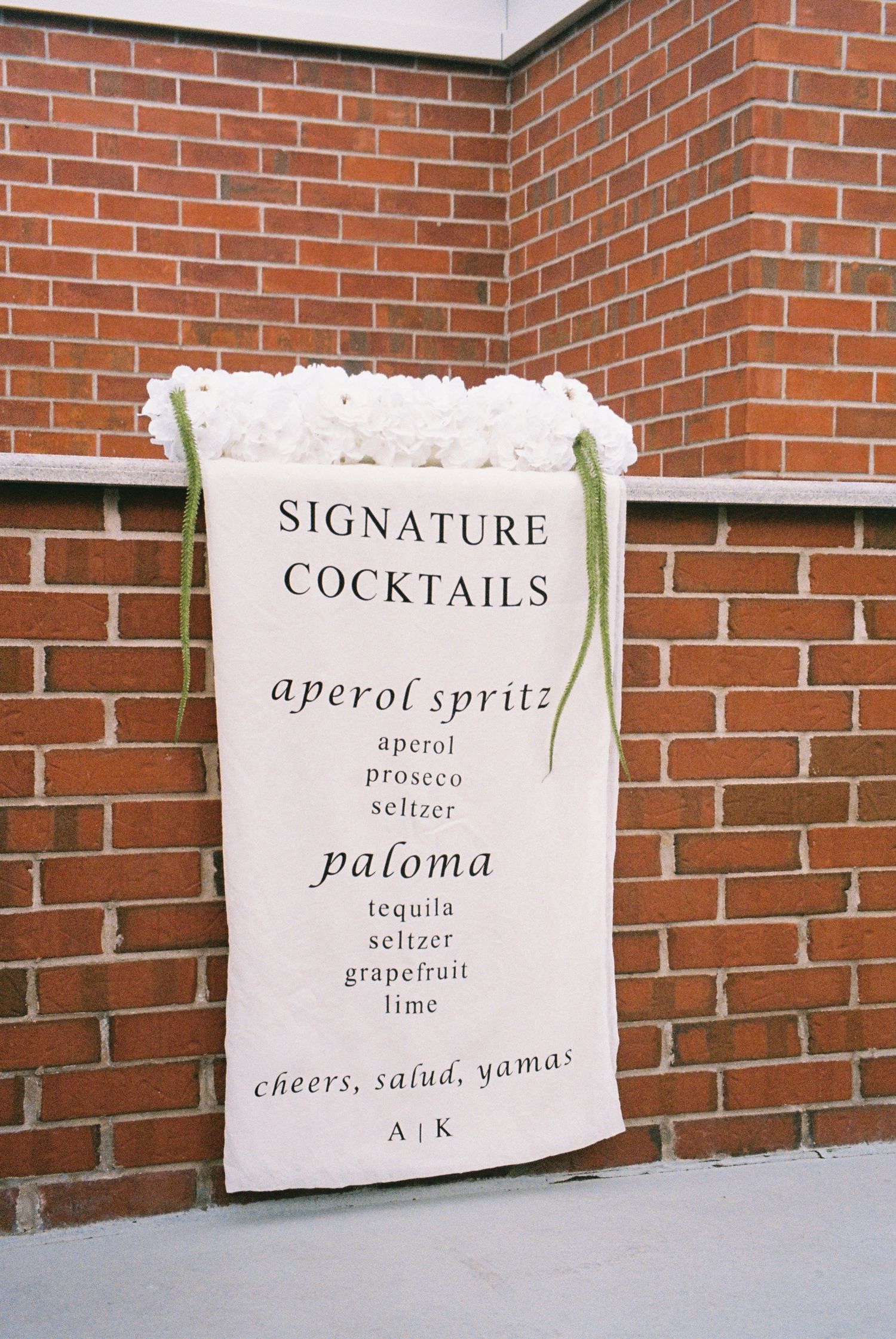 Signature cocktail menu printed on a large canvas for wedding reception guests to order from. It hangs over a red brick wall.