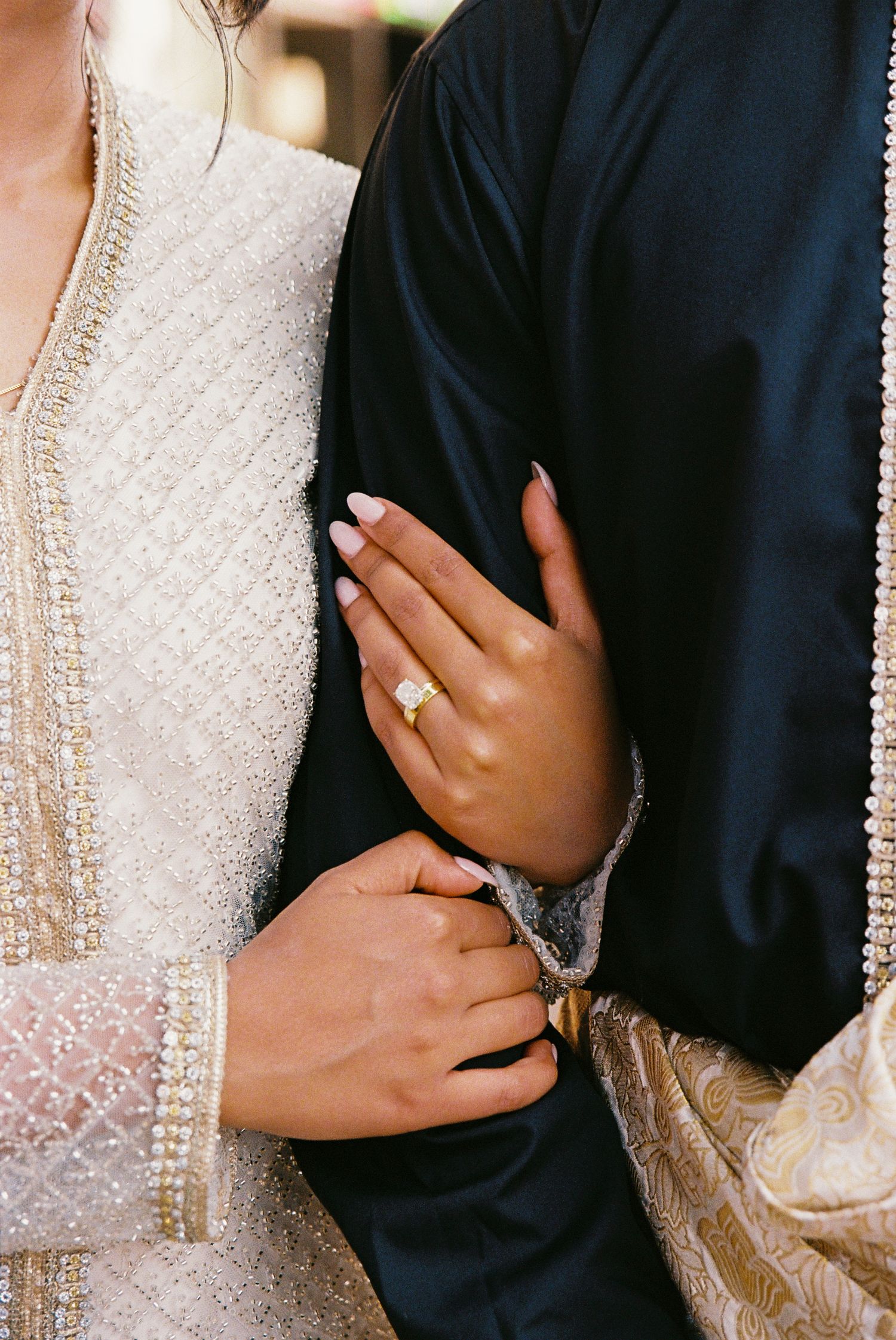 A wife's hands are wrapped around her husband's right arm. The focus of the film photo is the engagement ring on the bride's left hand.