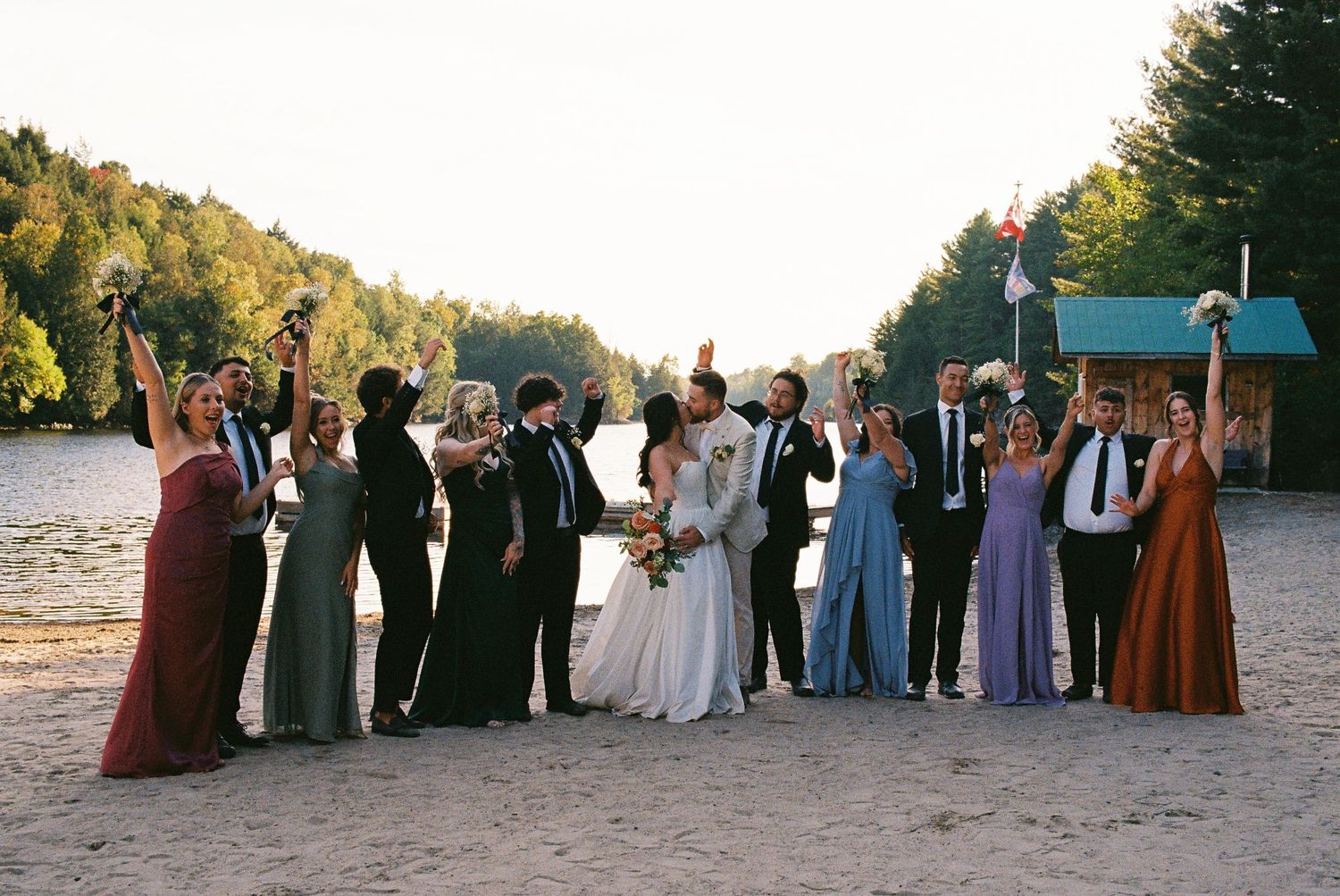 Bridesmaids and groomsmen posed in excitement next to the bride and groom kissing. The sun sets behind the subject.