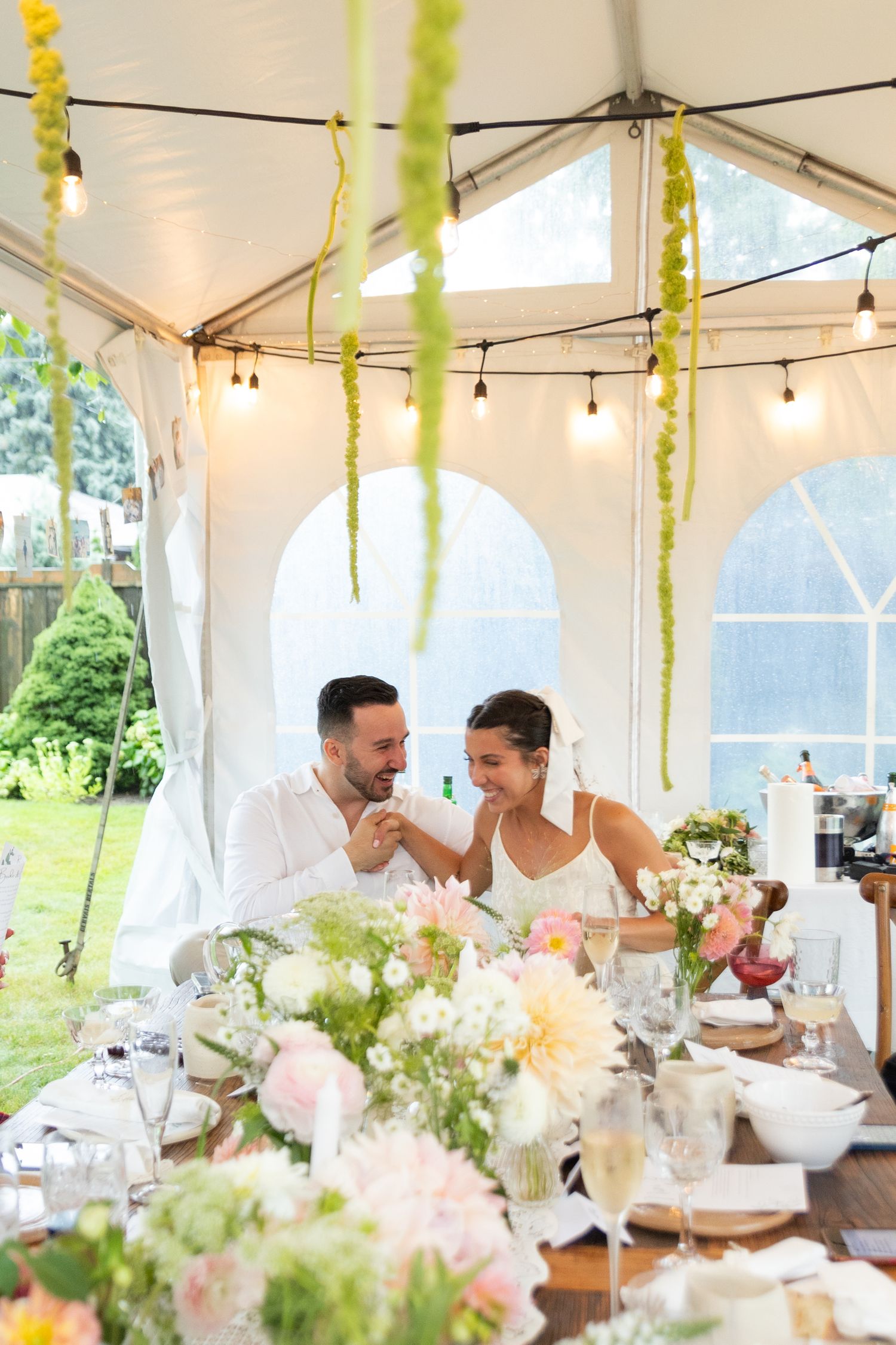 Bride and groom in backyard reception laughing and holding hands. The reception table is decorated in green, white, and light pink florals.