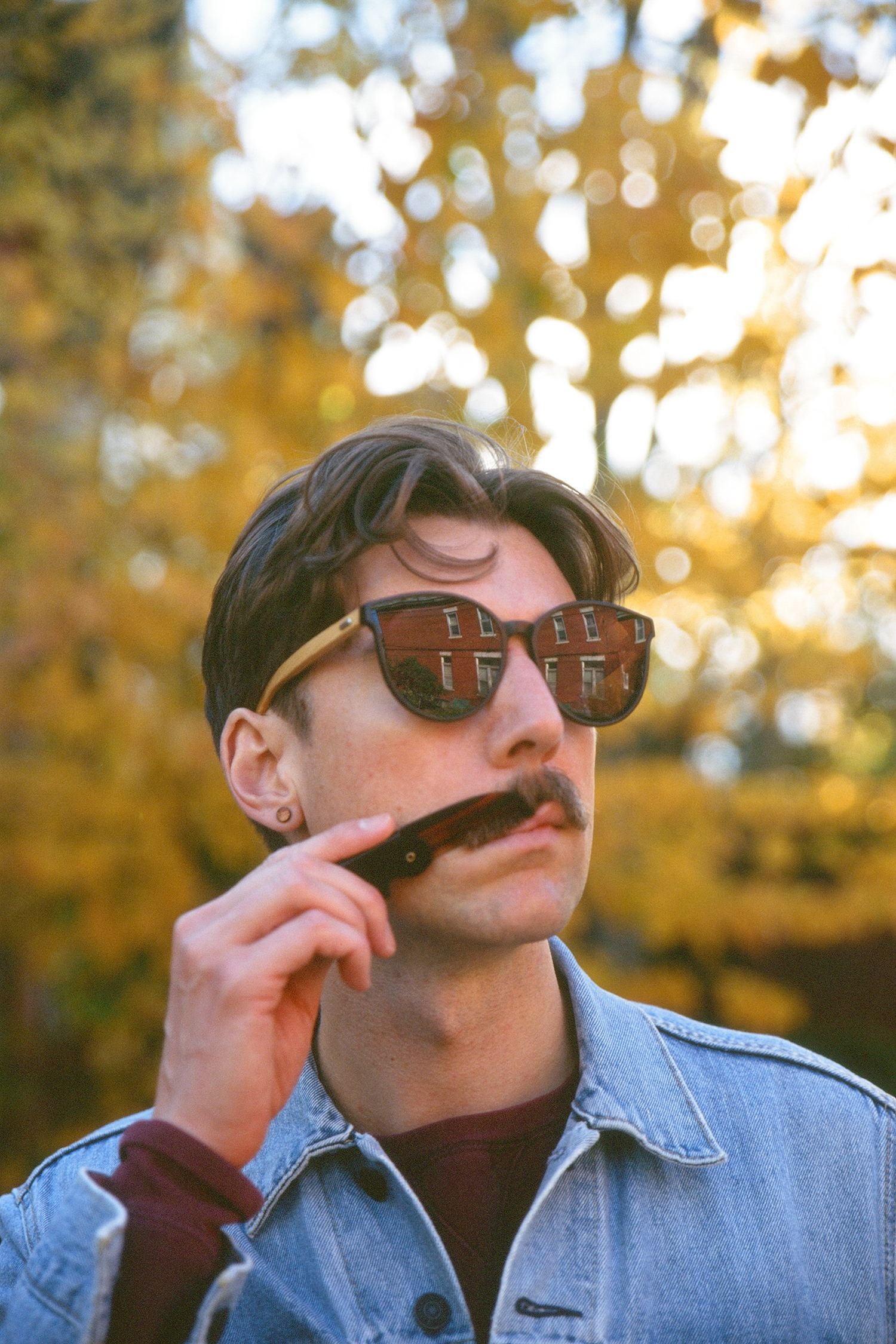 A man poses for a portrait photo wearing sunglasses and a denim shirt. He is combing his moustache in front of a tree with yellow leaves.
