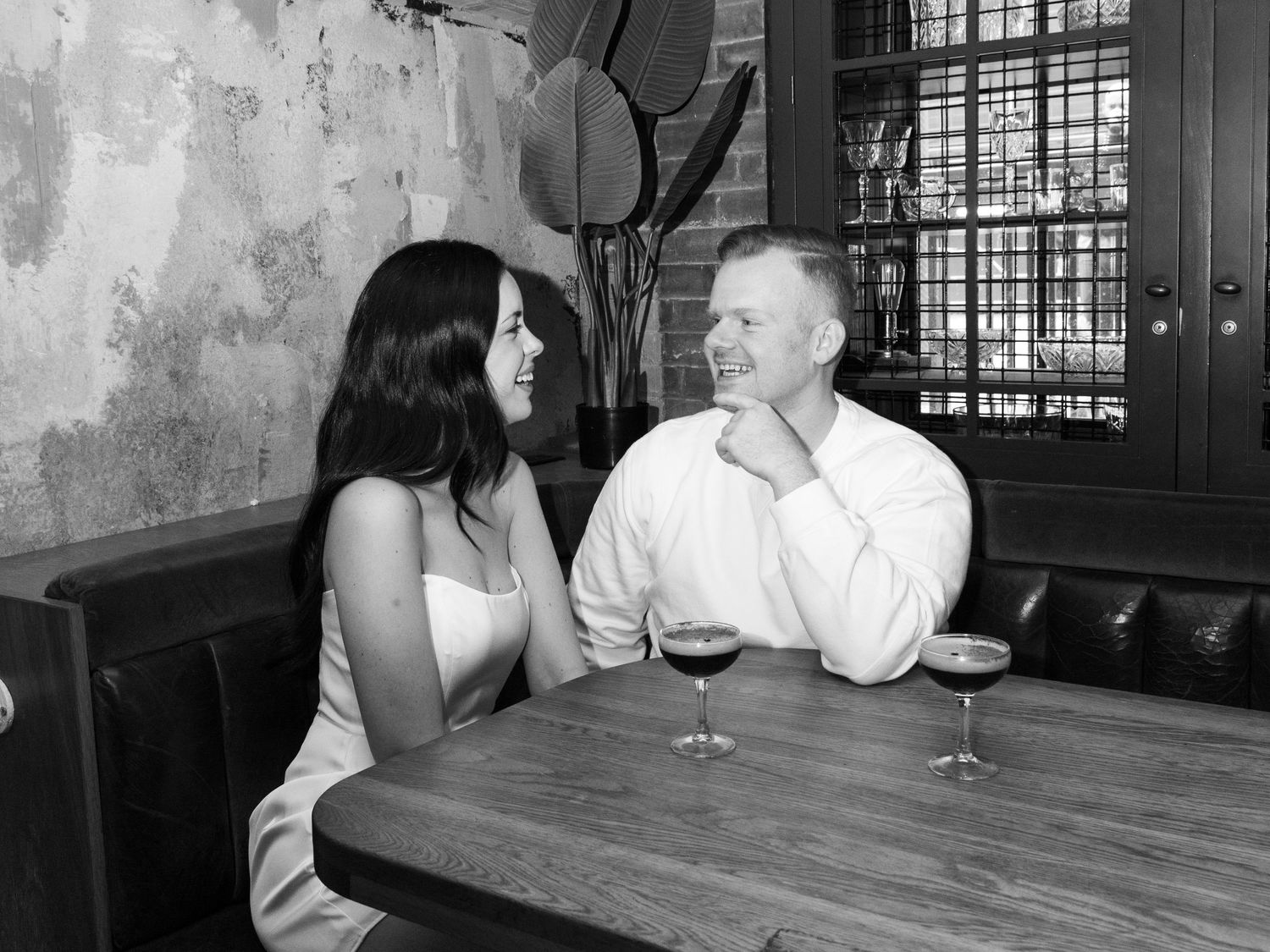 A couple sits together at a booth with espresso martinis and smile and laugh together.