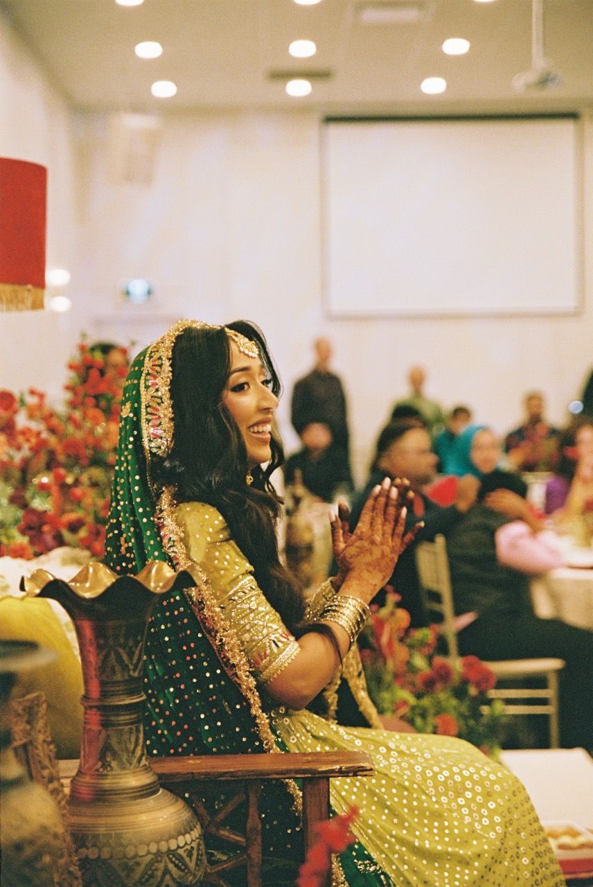 Bride is seated wearing shades of green at her mehndi and is smiling and clapping.