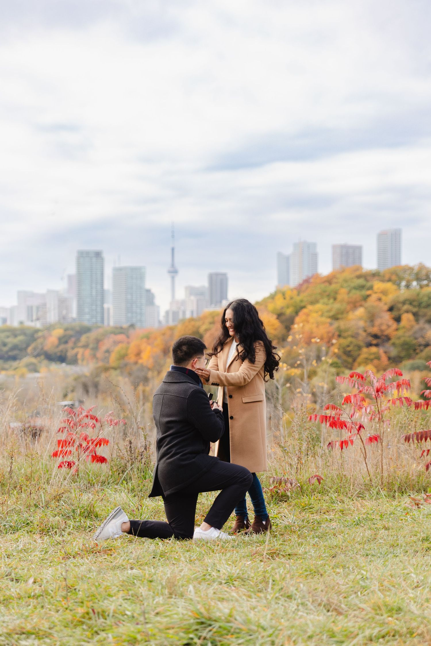 A man proposes to his girlfriend atop a hill at Evergreen Brickworks surrounded by fall leaves. The CN tower and Toronto skyline are behind.