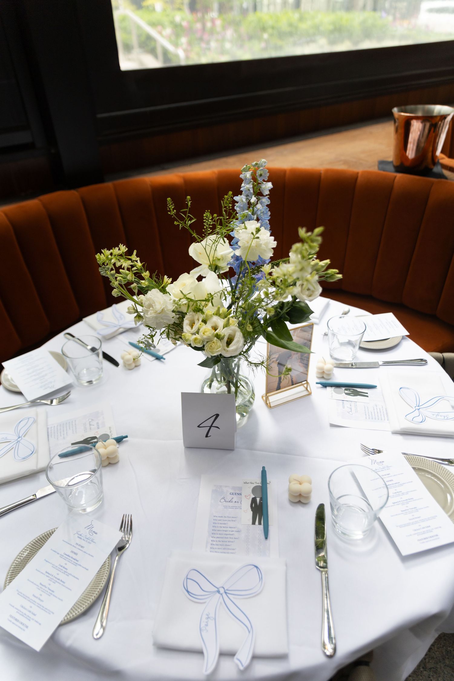 White table, brownish-orange booth bench, and floral decor set up at bridal shower. White menus with blue font cover the table.