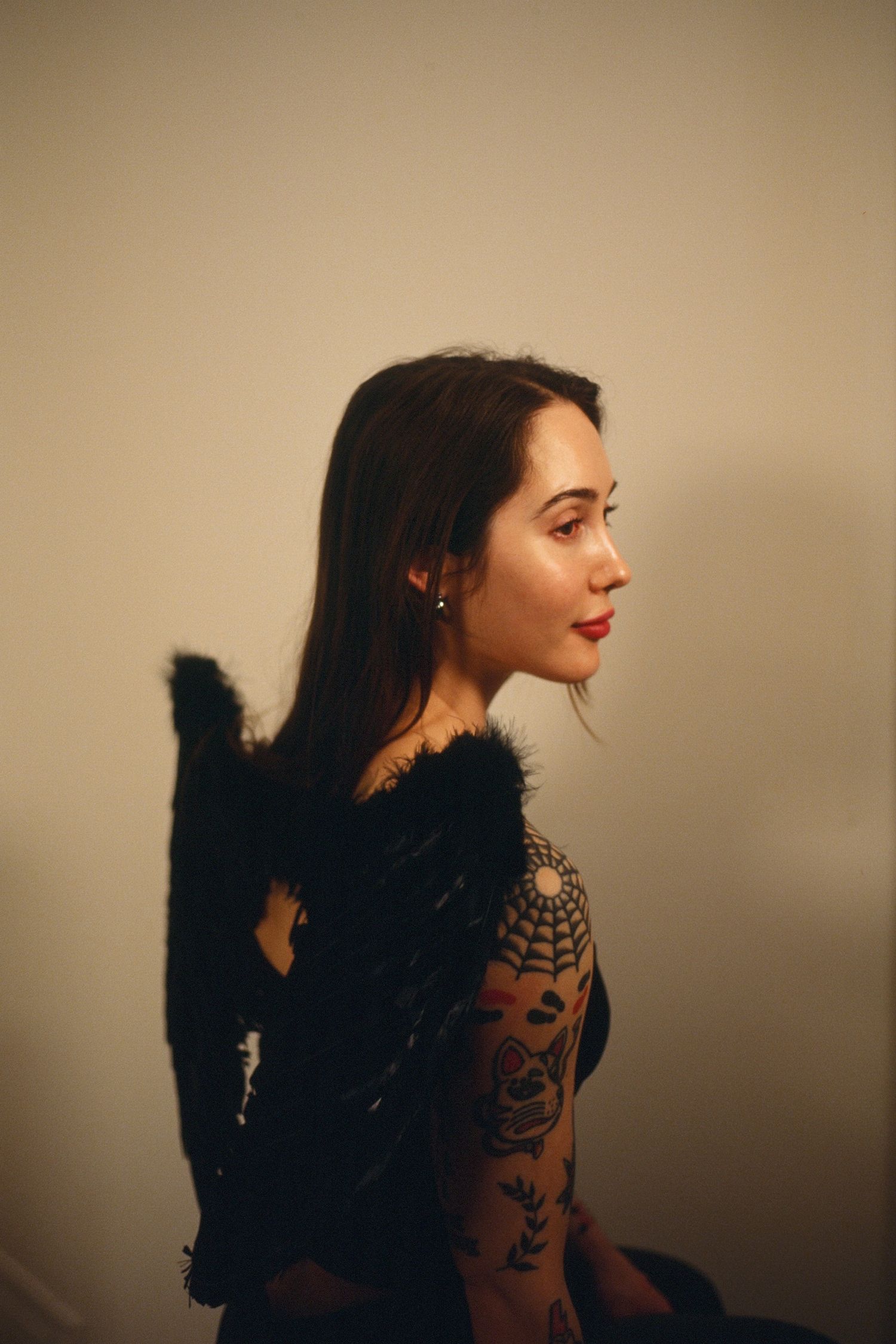 Film portrait of brown haired woman wearing black angel wings, posed in front of a white wall with ambient, warm lighting.