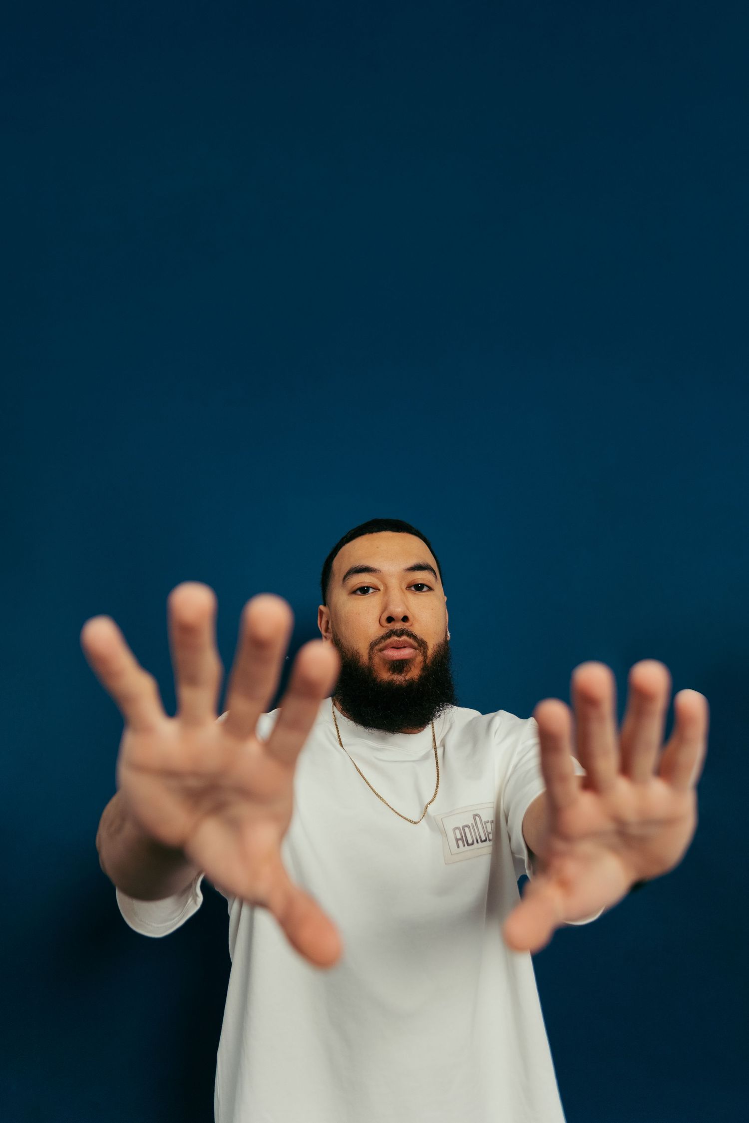 A man in a white t-shirt and gold necklace is posed in front of a blue backdrop with his hands up moving towards the camera.