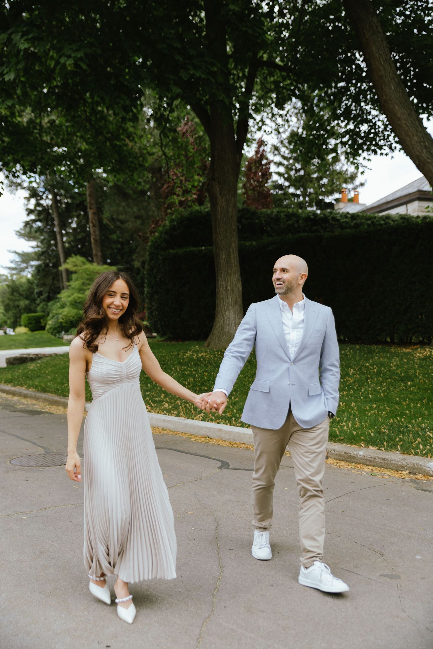 Two fiancés pose together on a quiet street lined with greenery before their engagement party.