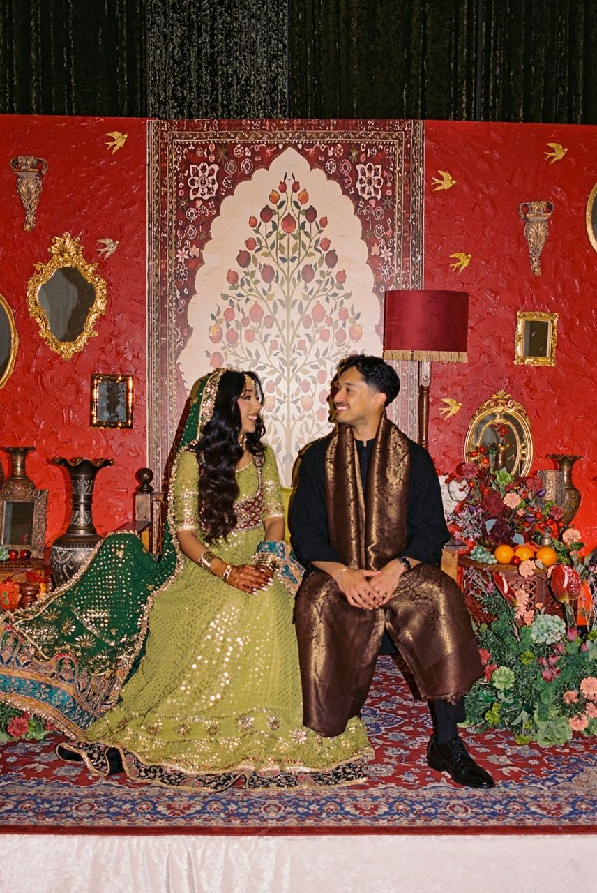 Bride and groom seated and smiling at each other in front of an intricate red, floral backdrop.