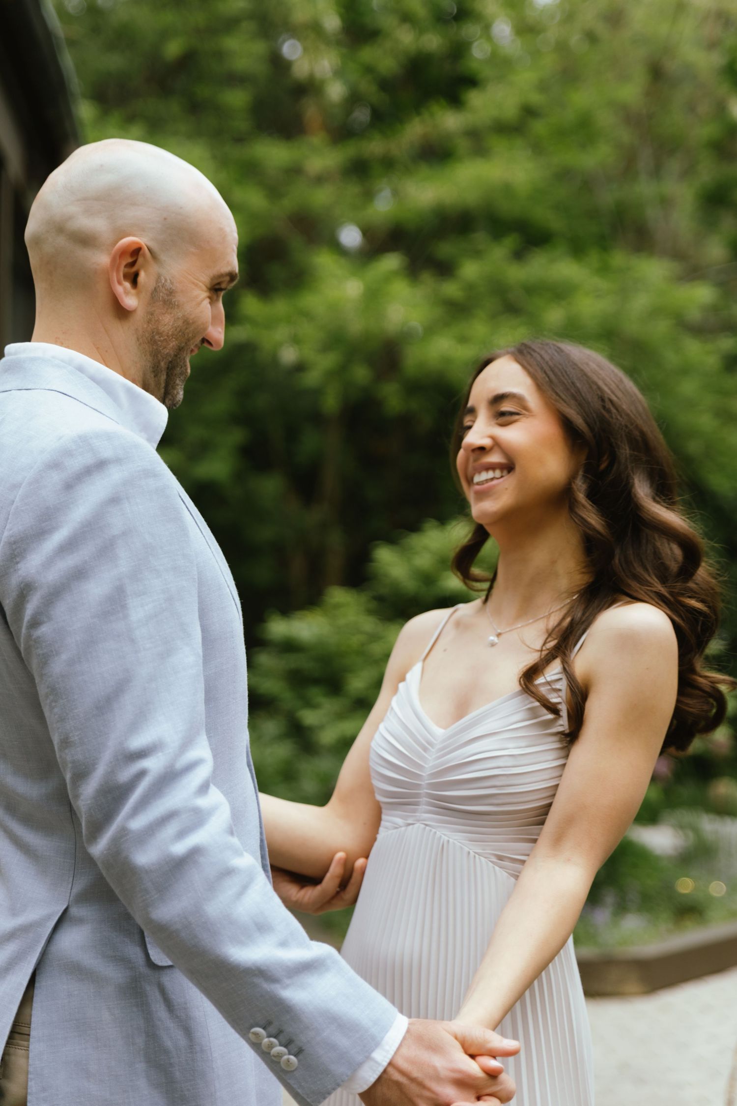 Two fiancés pose together on a quiet street lined with greenery before their engagement party. The bride-to-be is smiling and laughing.