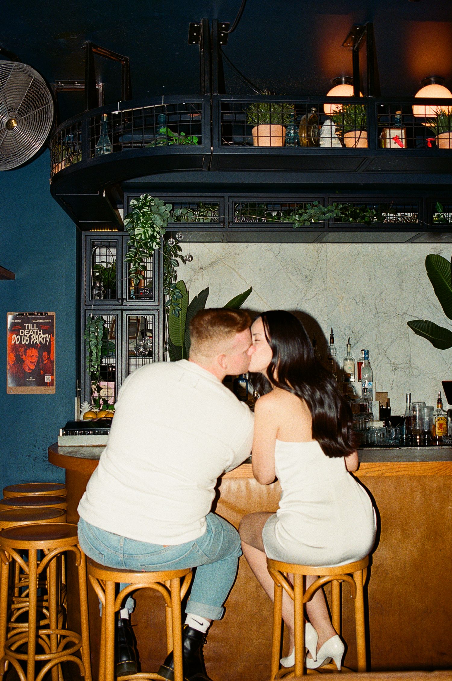 A couple dressed in white is seated at a bar and lean over to one another to kiss. Captured on 35mm film.