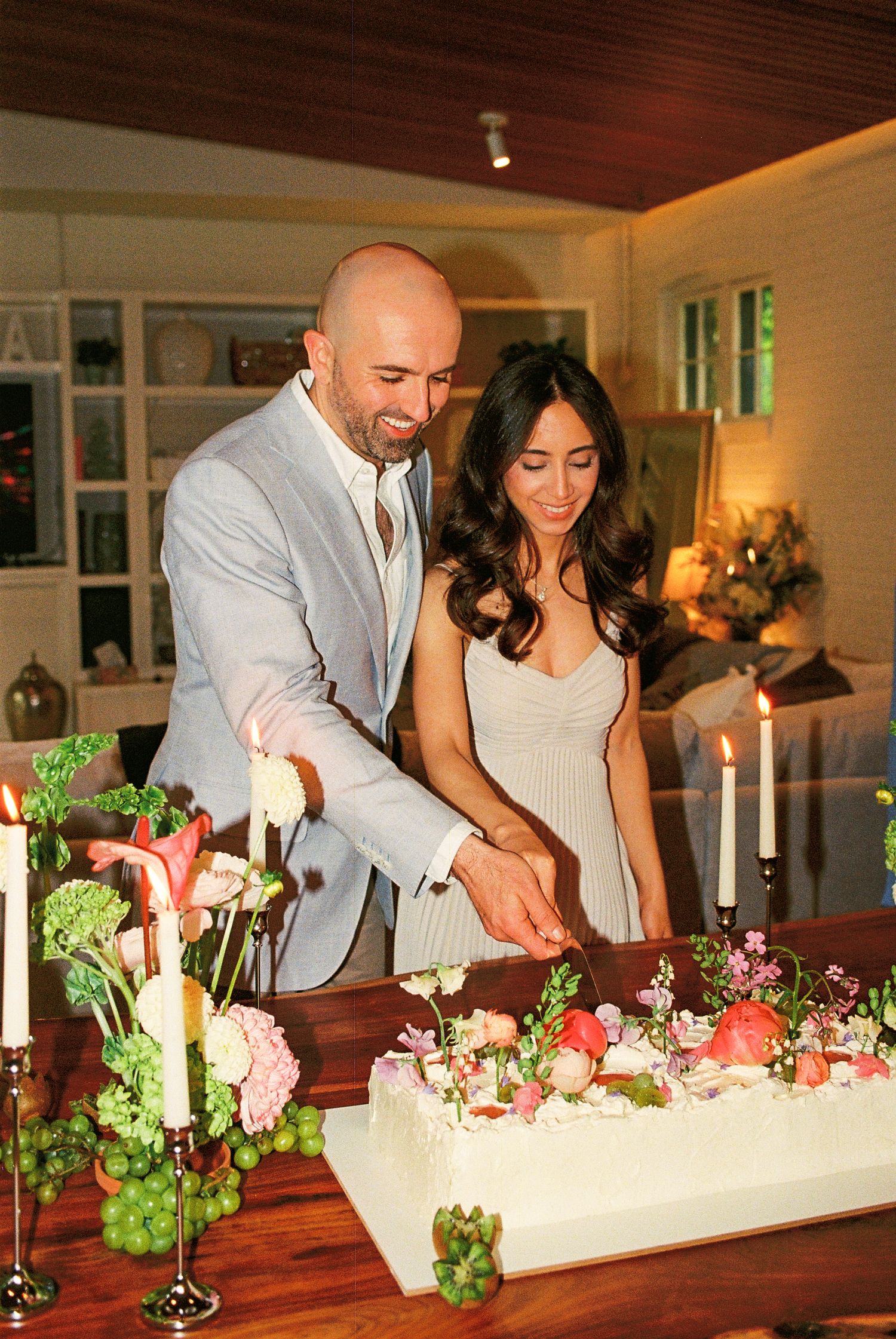 Fiancés smiling and holding hands while cutting their white, green, and pink floral cake at their engagement party.