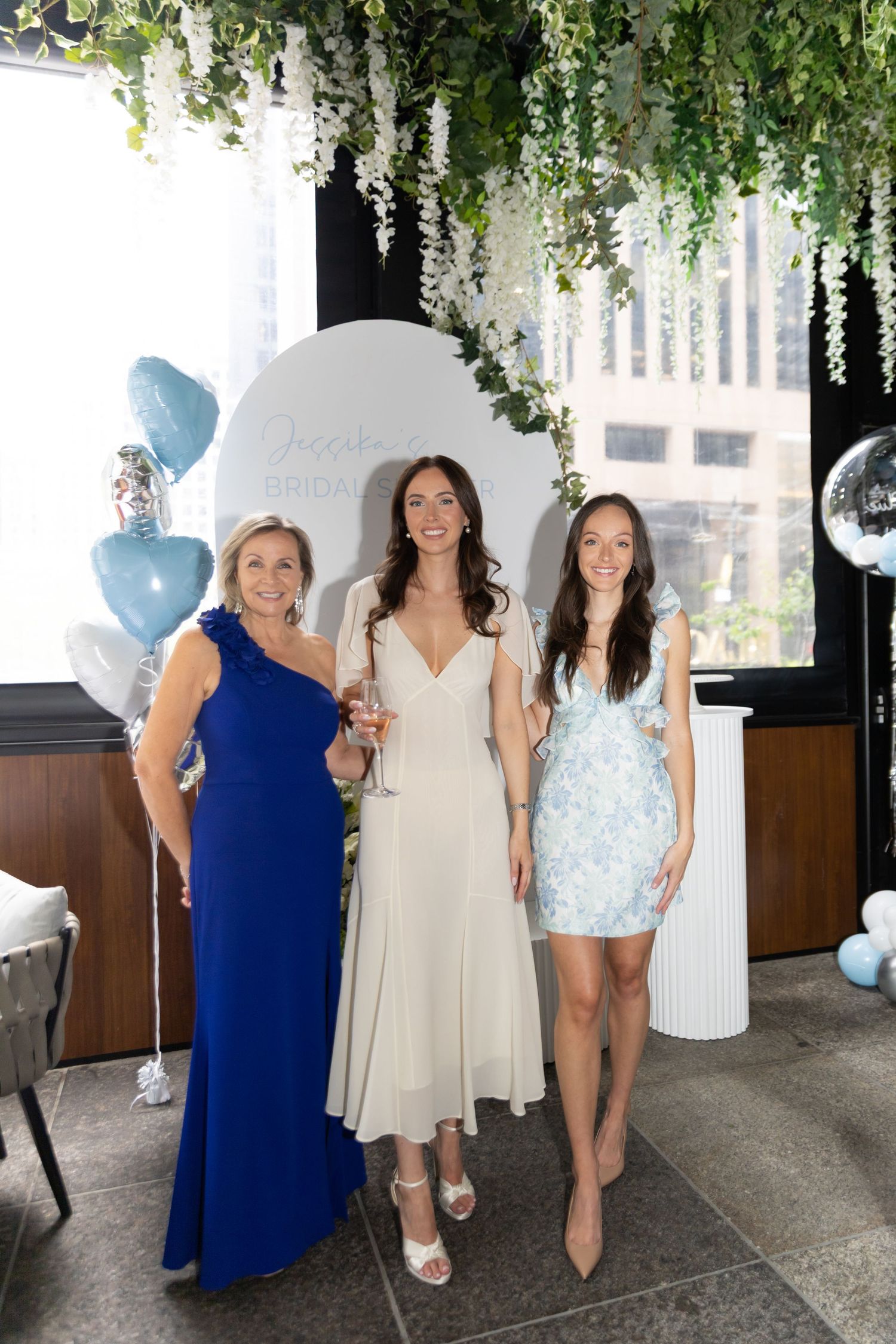 Group photo of bride, mother, and sister at bridal shower. Greenery lines the ceiling of the venue