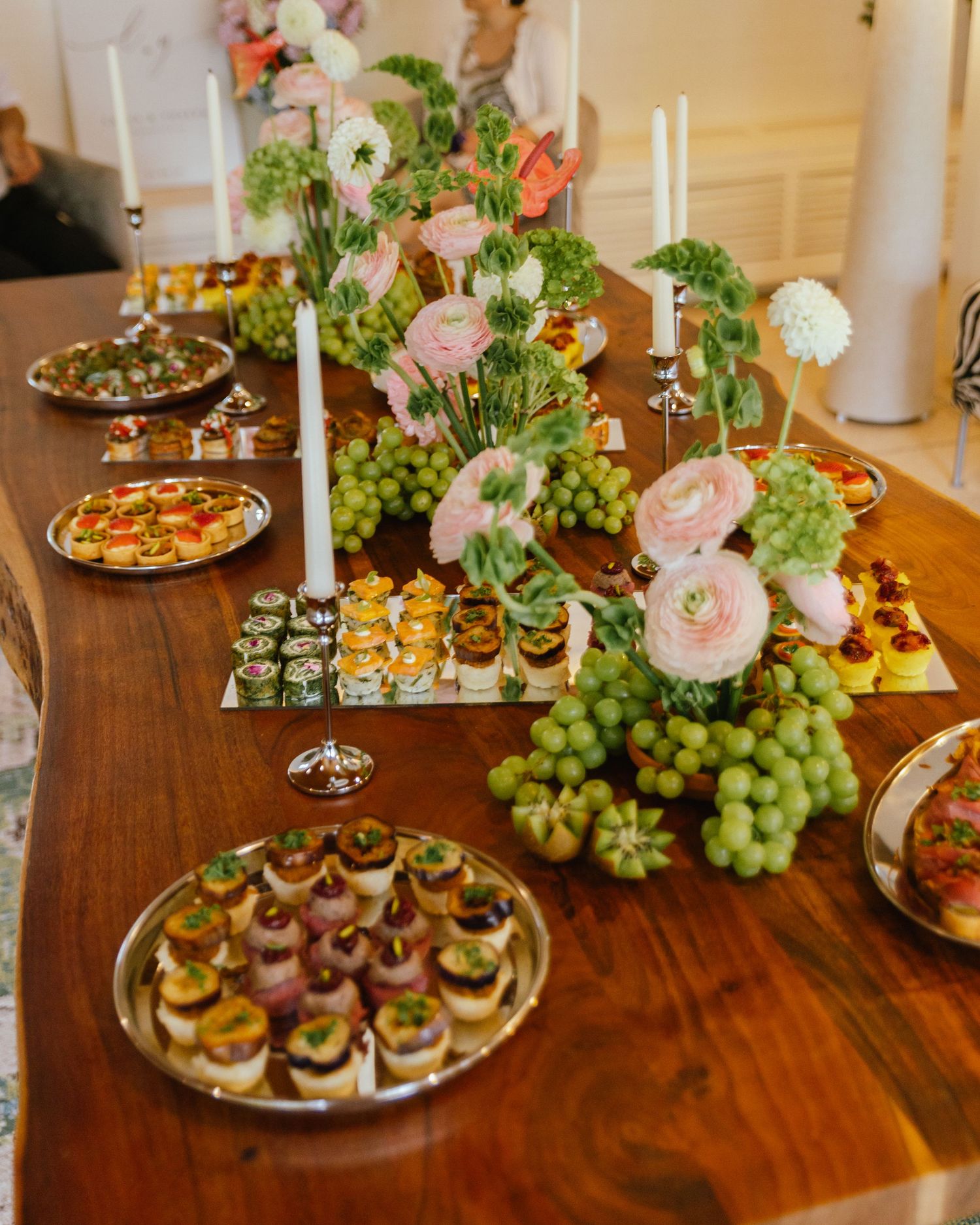 Wooden table filled with green and pink flowers, tapered candles, and hors d'ouevres at an engagement party.