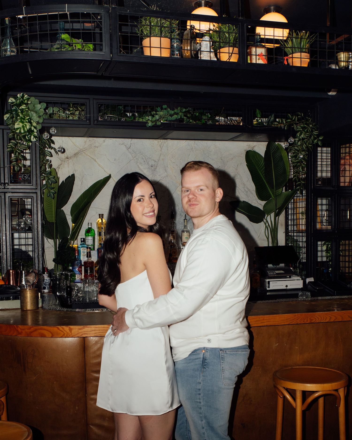 Fiancés posed together a bar where they met in Toronto. They are facing the bar and looking over their shoulders to smile at the camera.