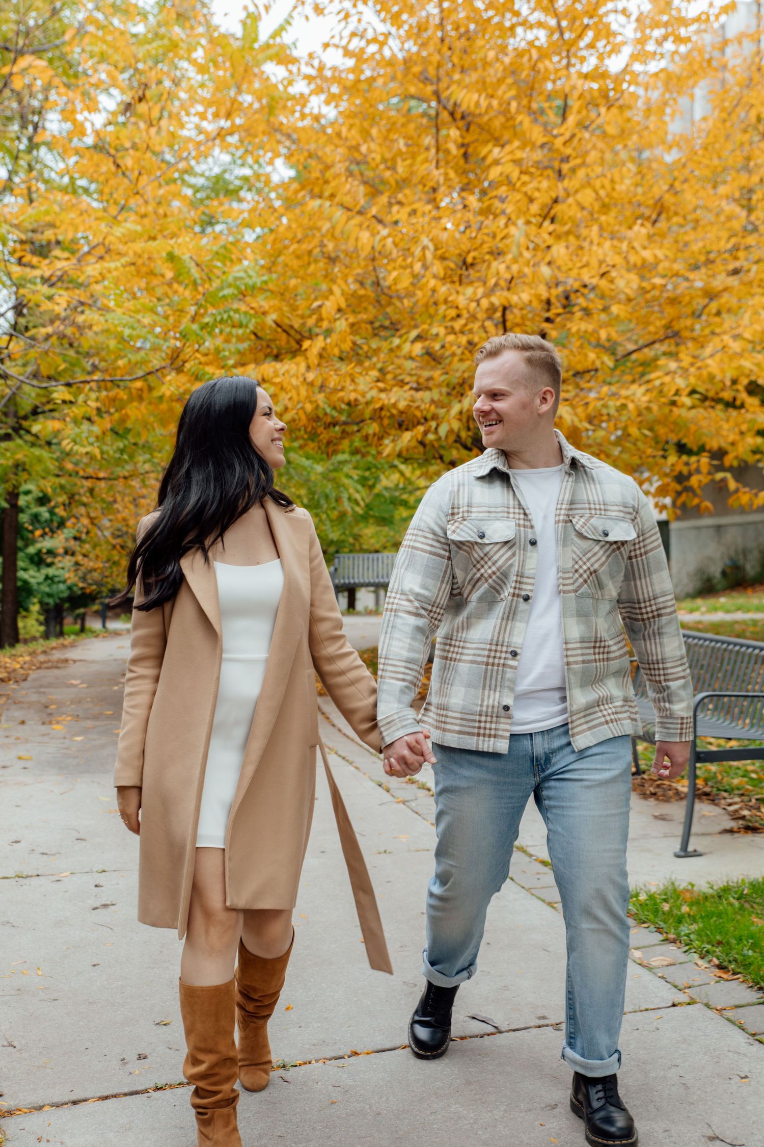 Fiancés holding hands walking towards the camera while smiling and looking into each other's eyes. They are surrounded by fall foliage.
