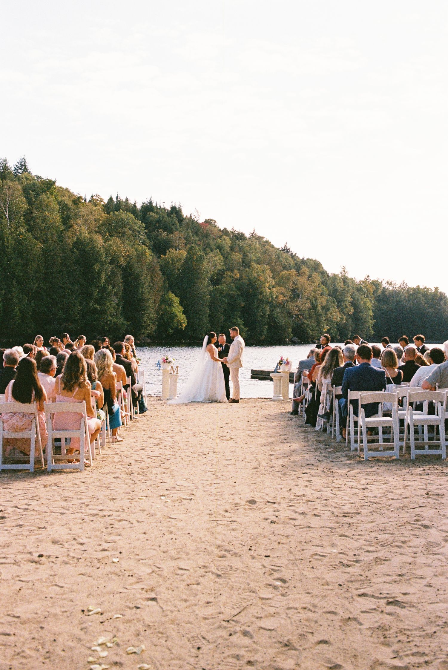 Wedding ceremony on a beach captured on film, the bride and groom are centre aisle and rows of guests seated in white chairs on each side.