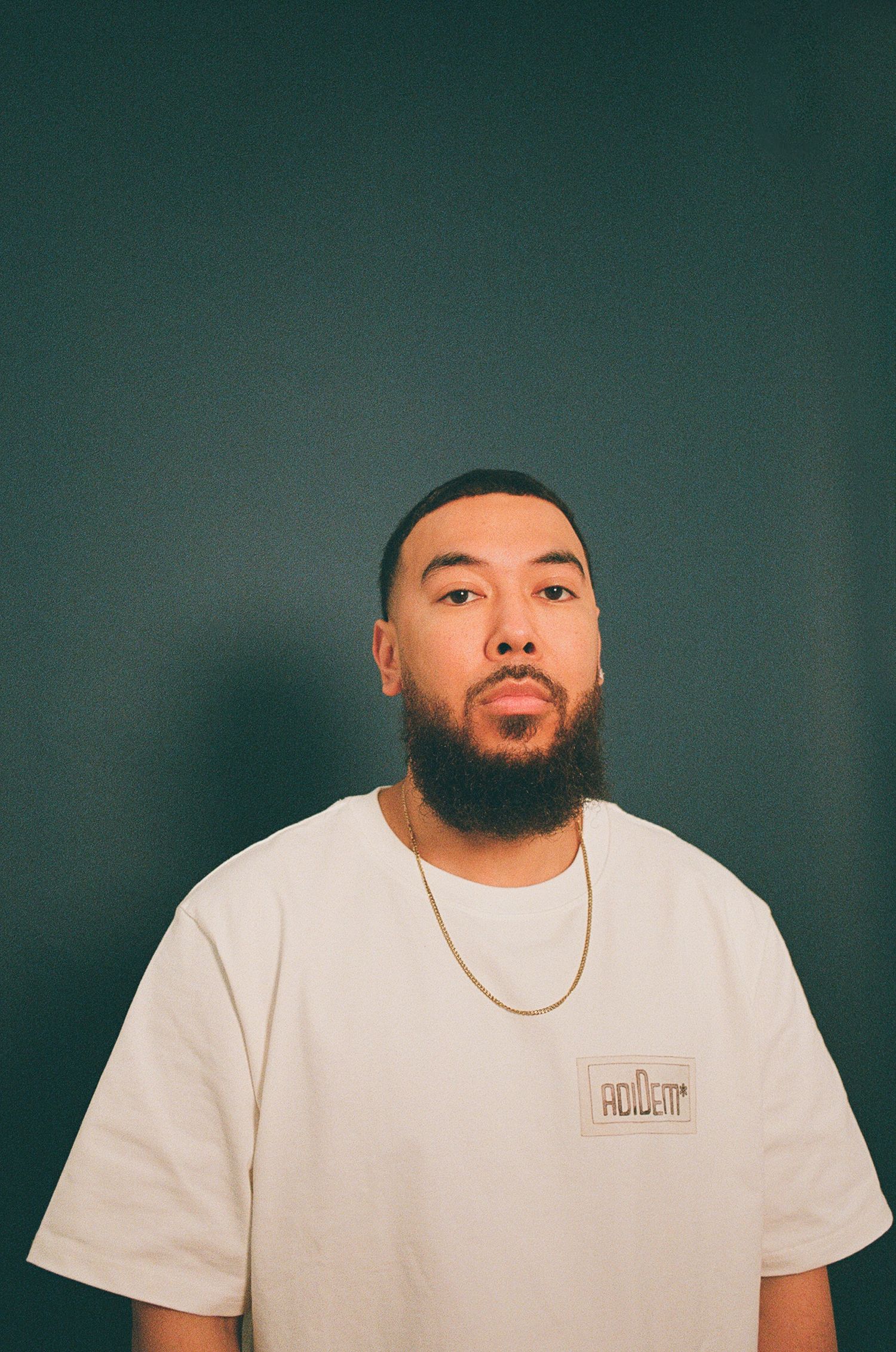 A man wearing a white t-shirt poses for a film photo in front of a teal coloured studio backdrop.