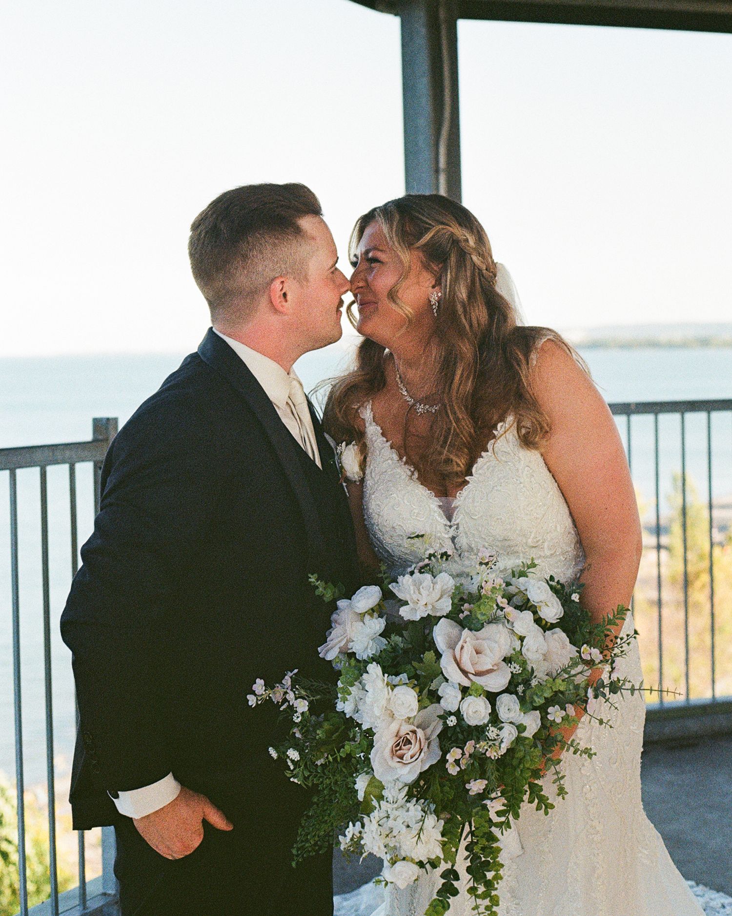 Bride and groom touching noses and smiling. The bride is holding a lush bouquet of flowers and greenery and blurred behind them is a lake.