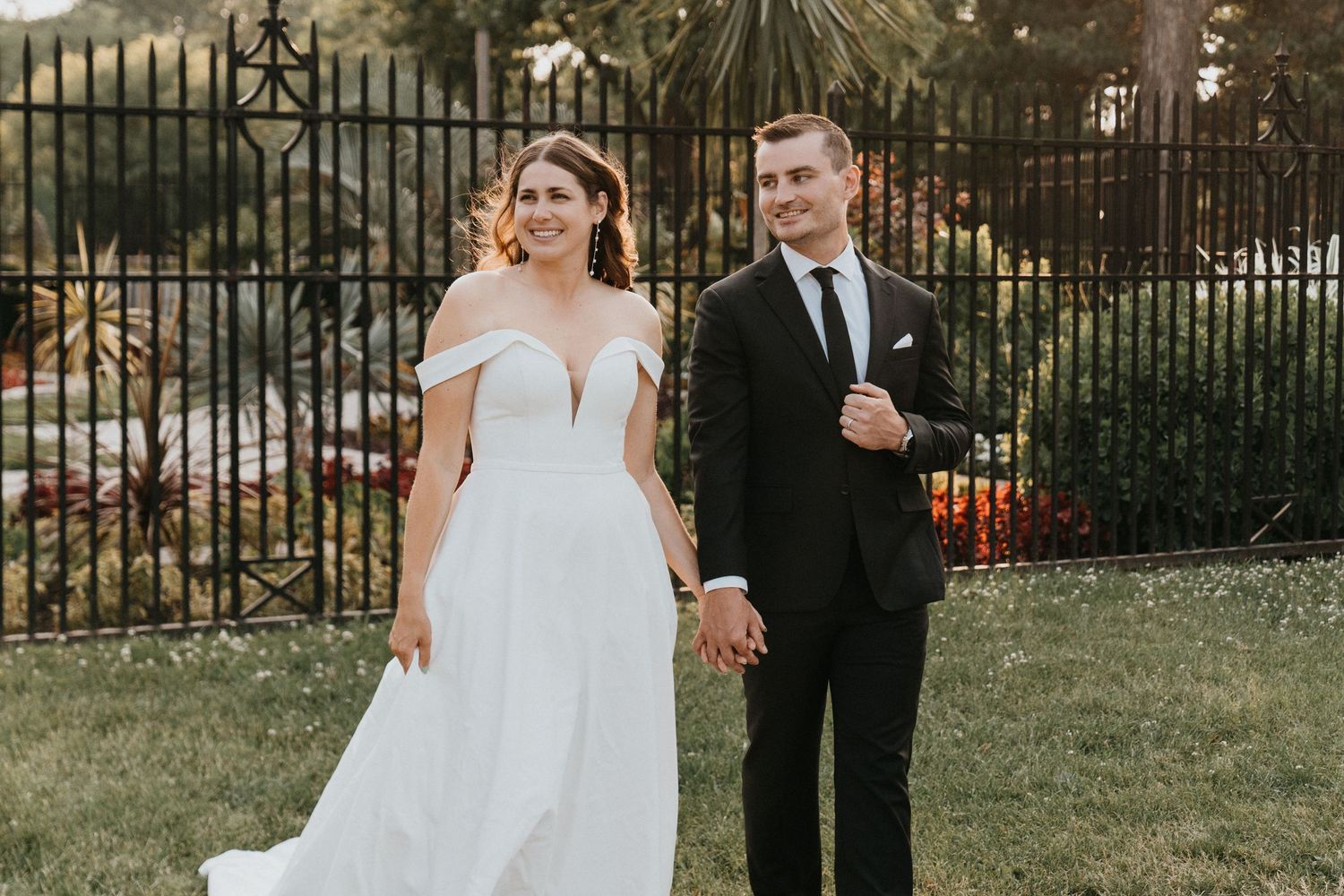 A couple in wedding attire poses elegantly against an ornate iron gate and garden backdrop.