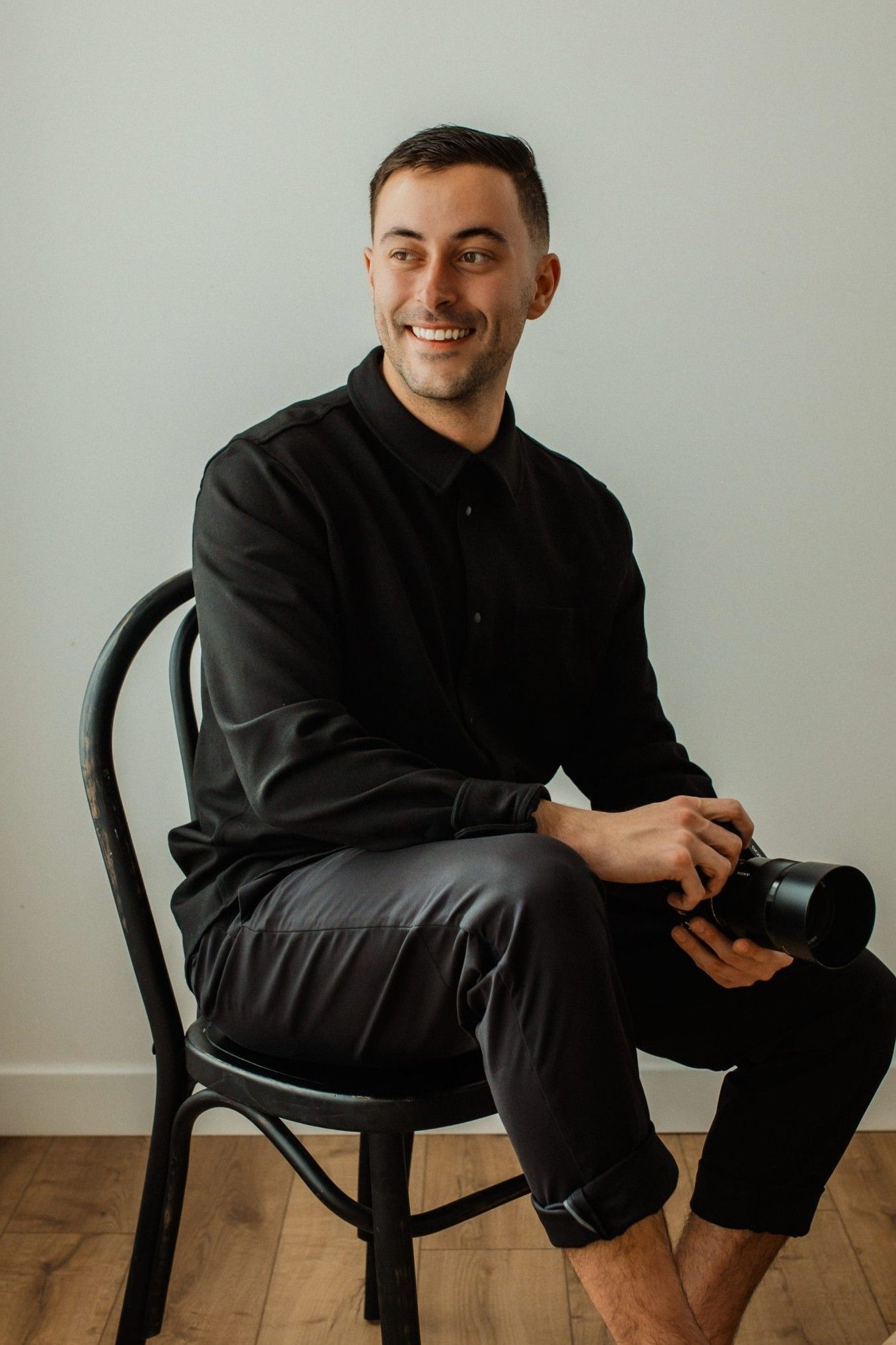 Person in black clothing sits casually on a chair against a minimalist gray background.