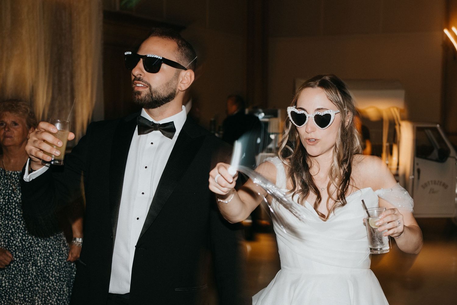Wedding party guests in formal attire holding drinks and wearing sunglasses at an elegant reception.