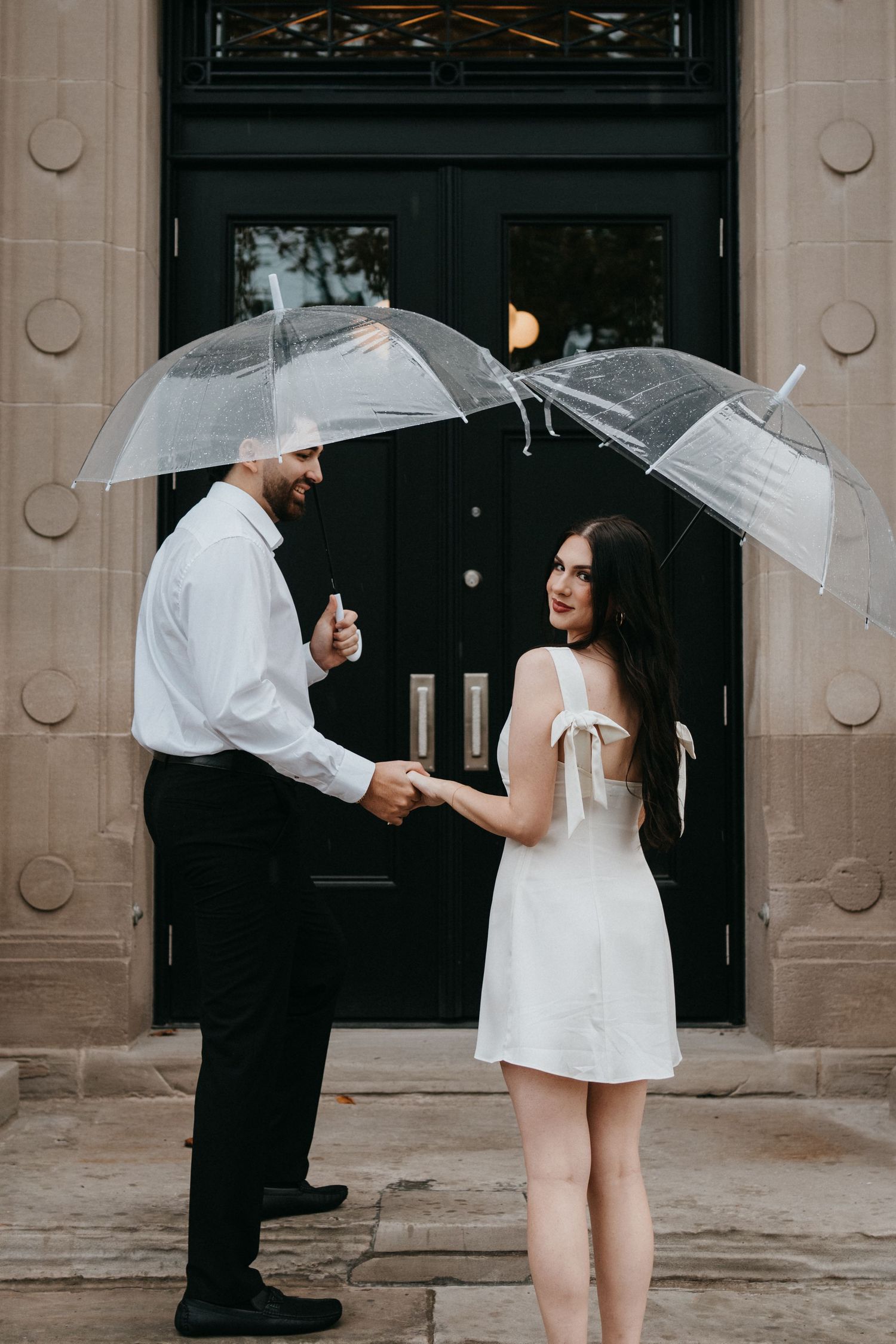 A couple in formal attire holding clear umbrellas stand in front of a dark doorway on a stone building.