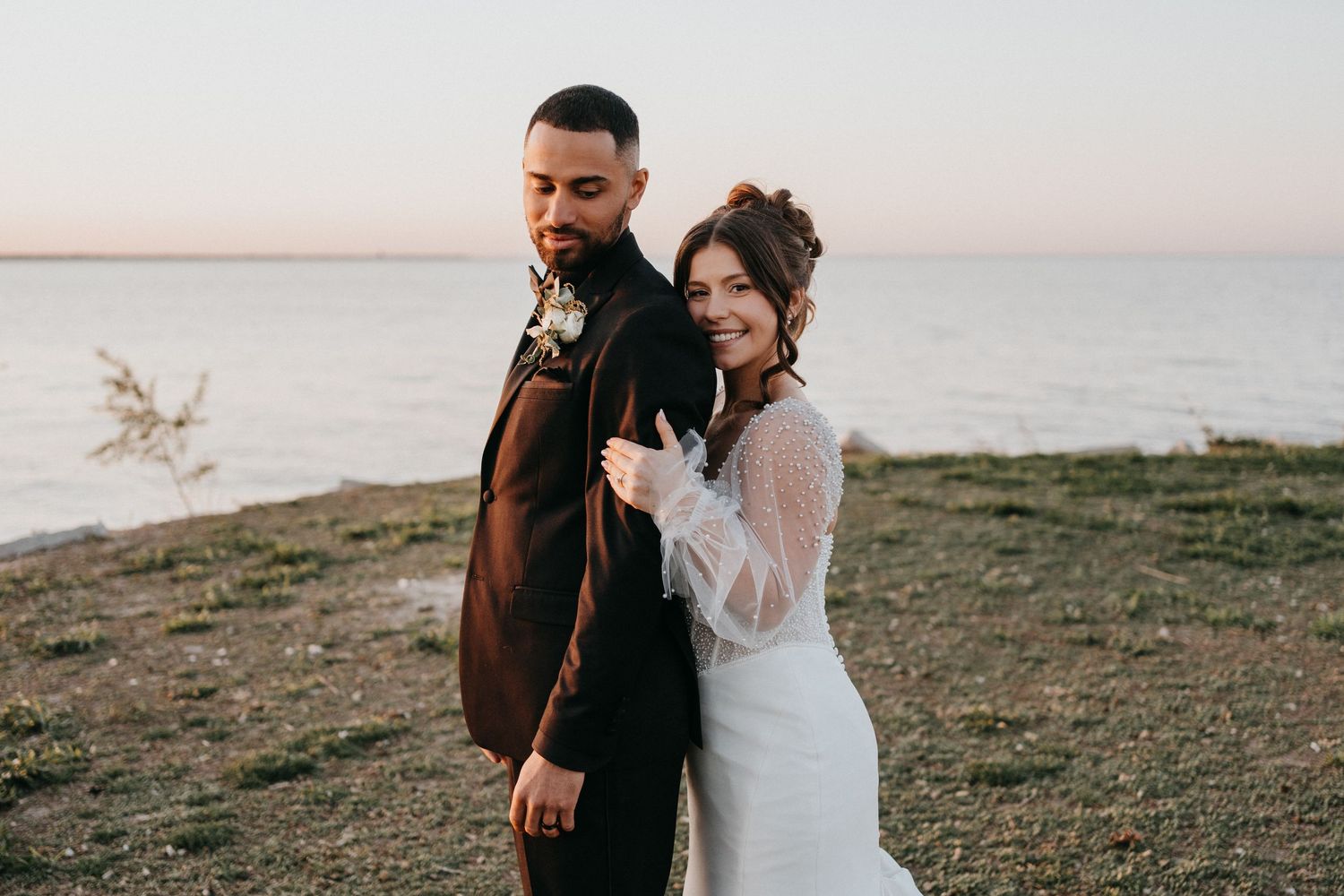 A couple shares an intimate moment during their wedding photoshoot at sunset on a grassy coastal cliff.