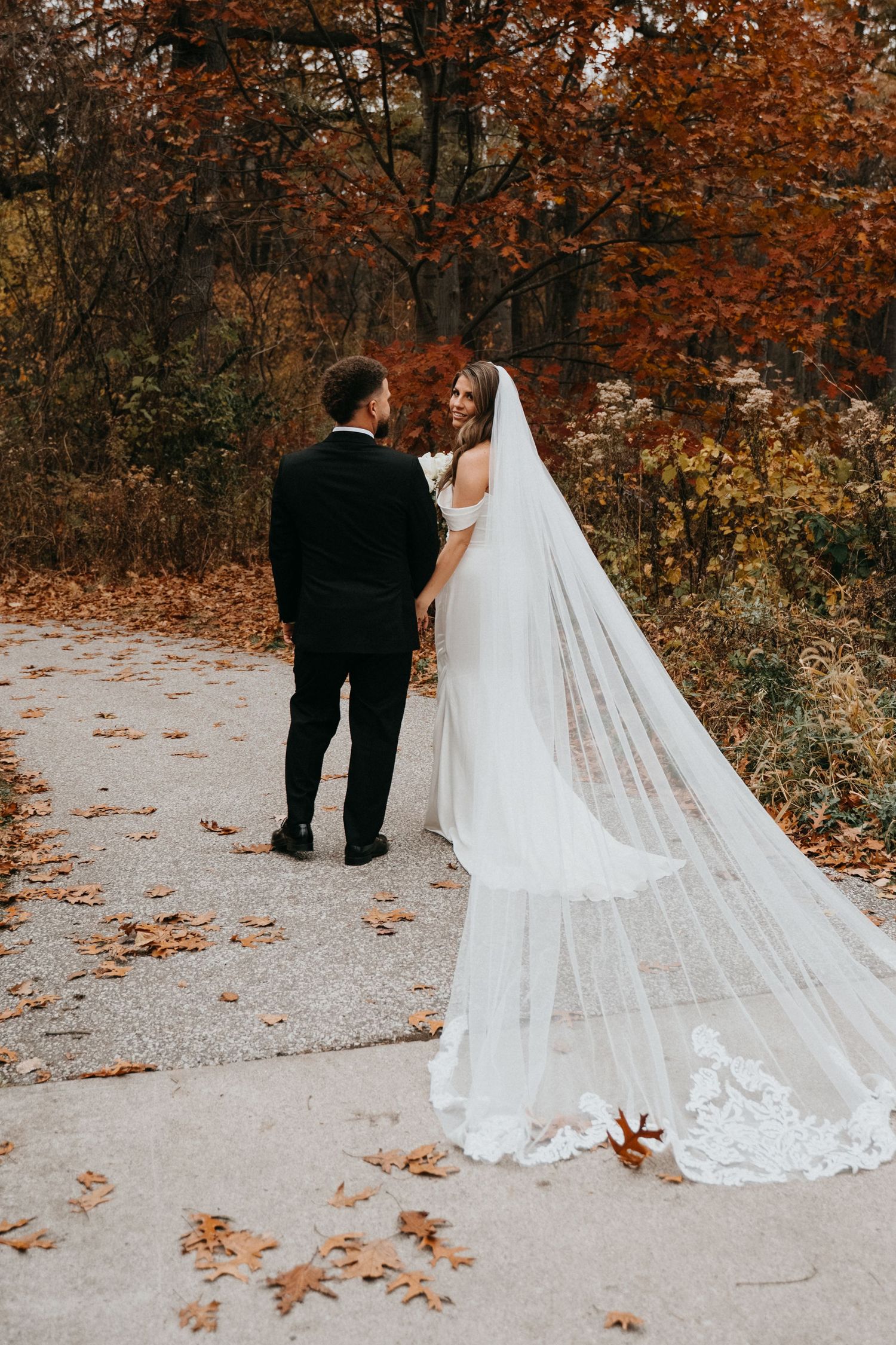 Wedding couple walking on autumn path with long lace veil trailing behind them.