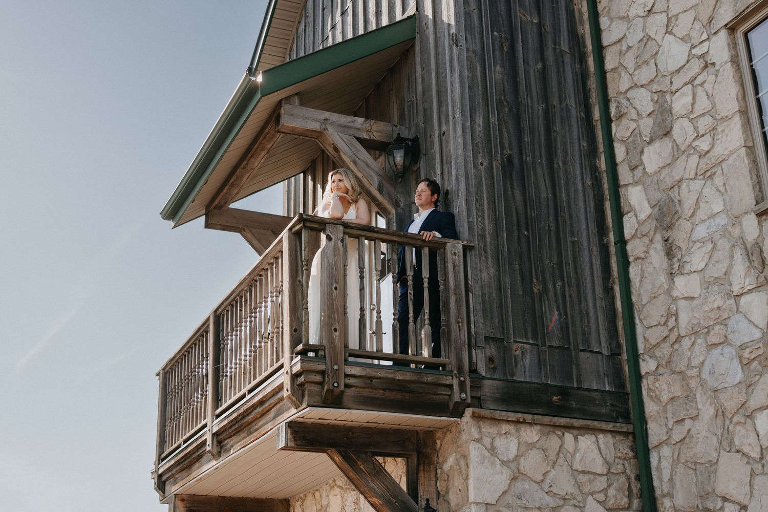 Couple in wedding attire standing on a rustic wooden balcony of a stone building against a clear sky.