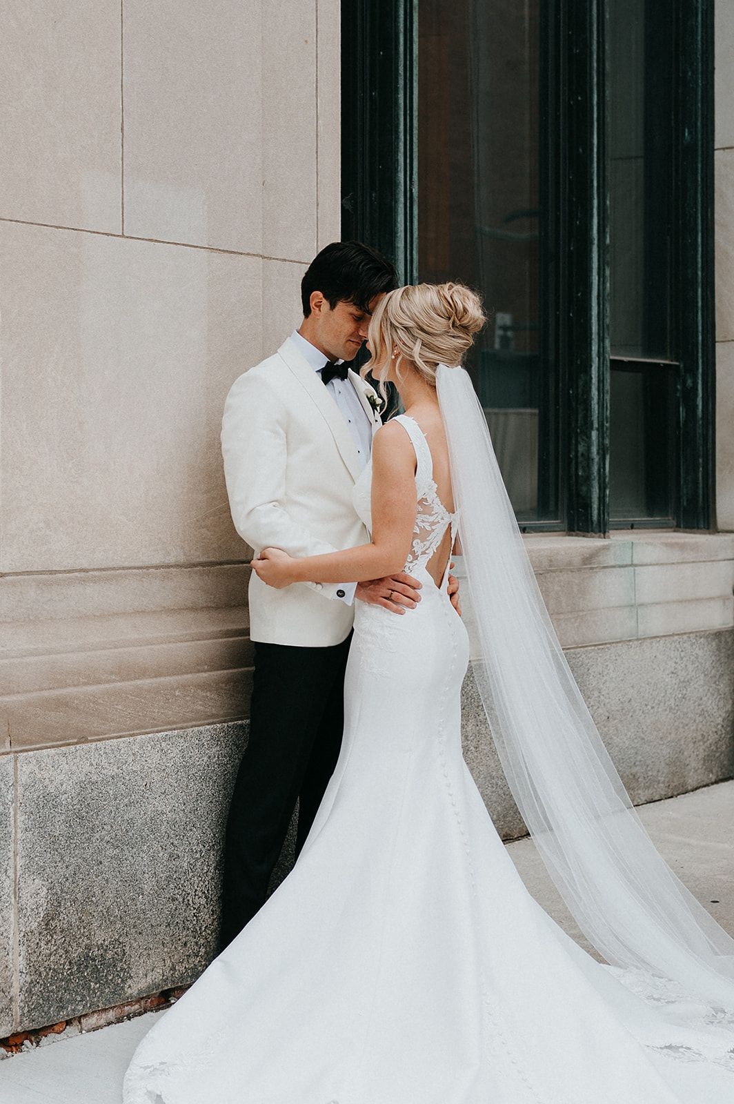 Bride in lace mermaid gown with veil and groom in white tuxedo jacket share romantic moment against stone building.