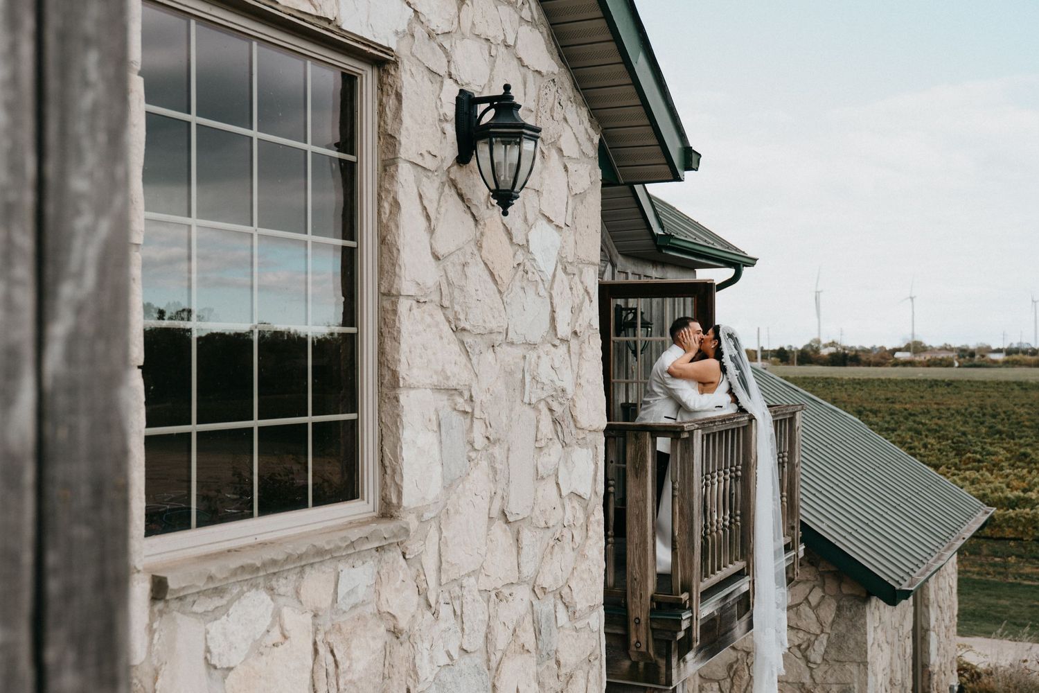Wedding couple poses outside rustic stone building with large windows.