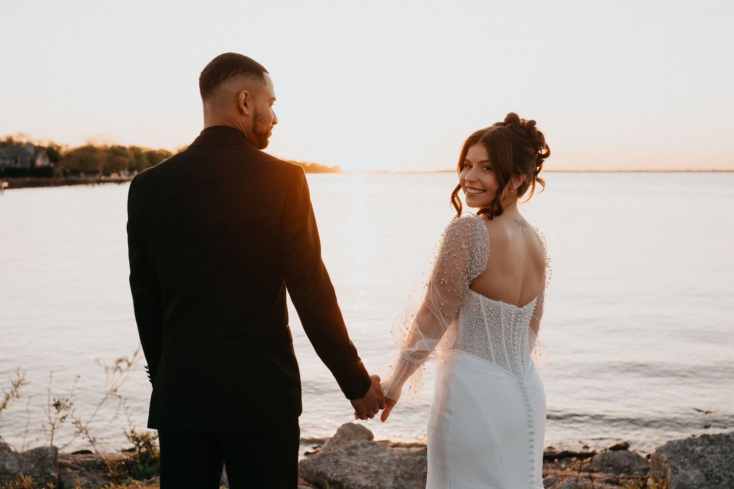 Newlyweds share intimate moment at sunset by waterfront.