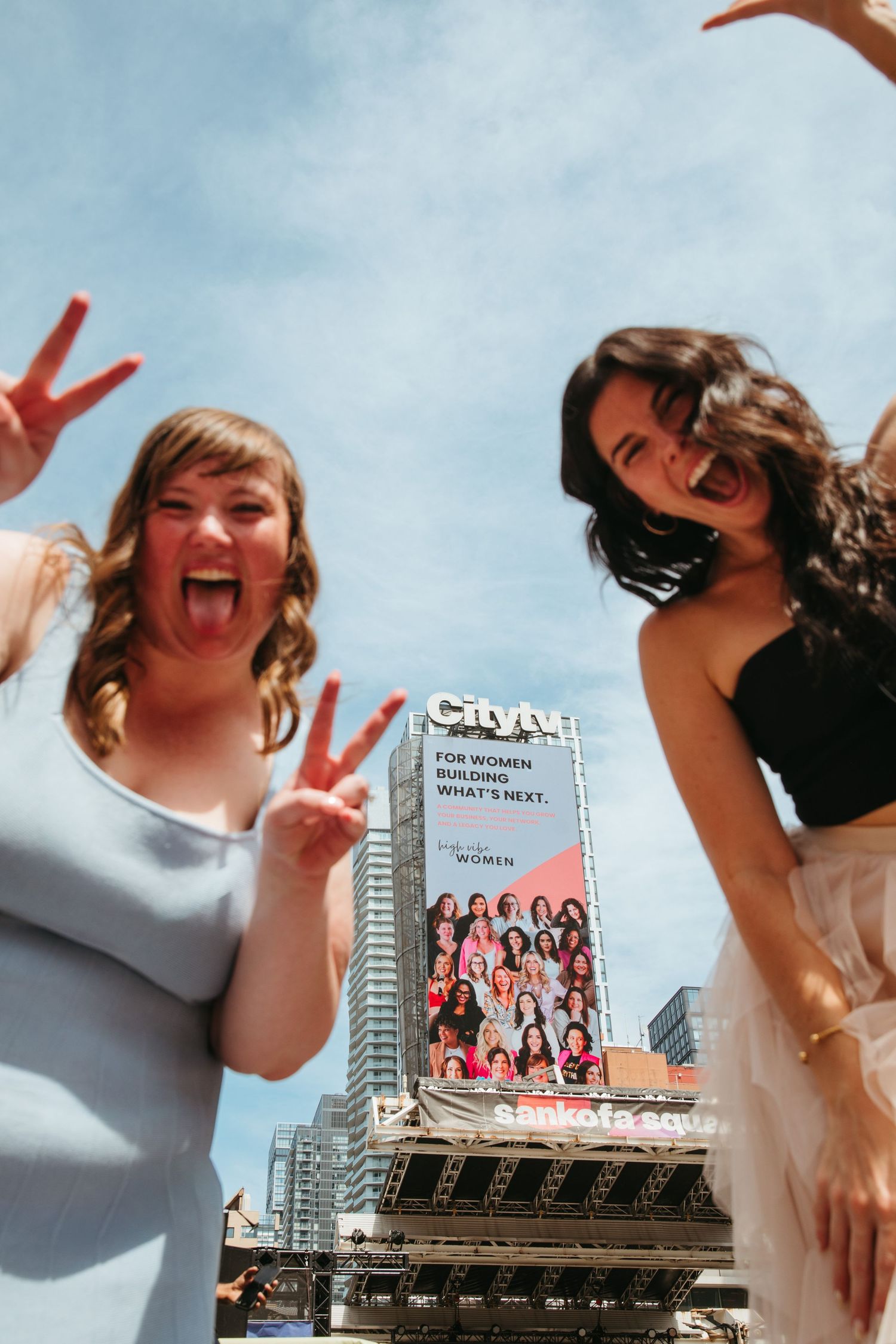 Two women posing with peace signs in front of a CityTV billboard for women in an urban setting.