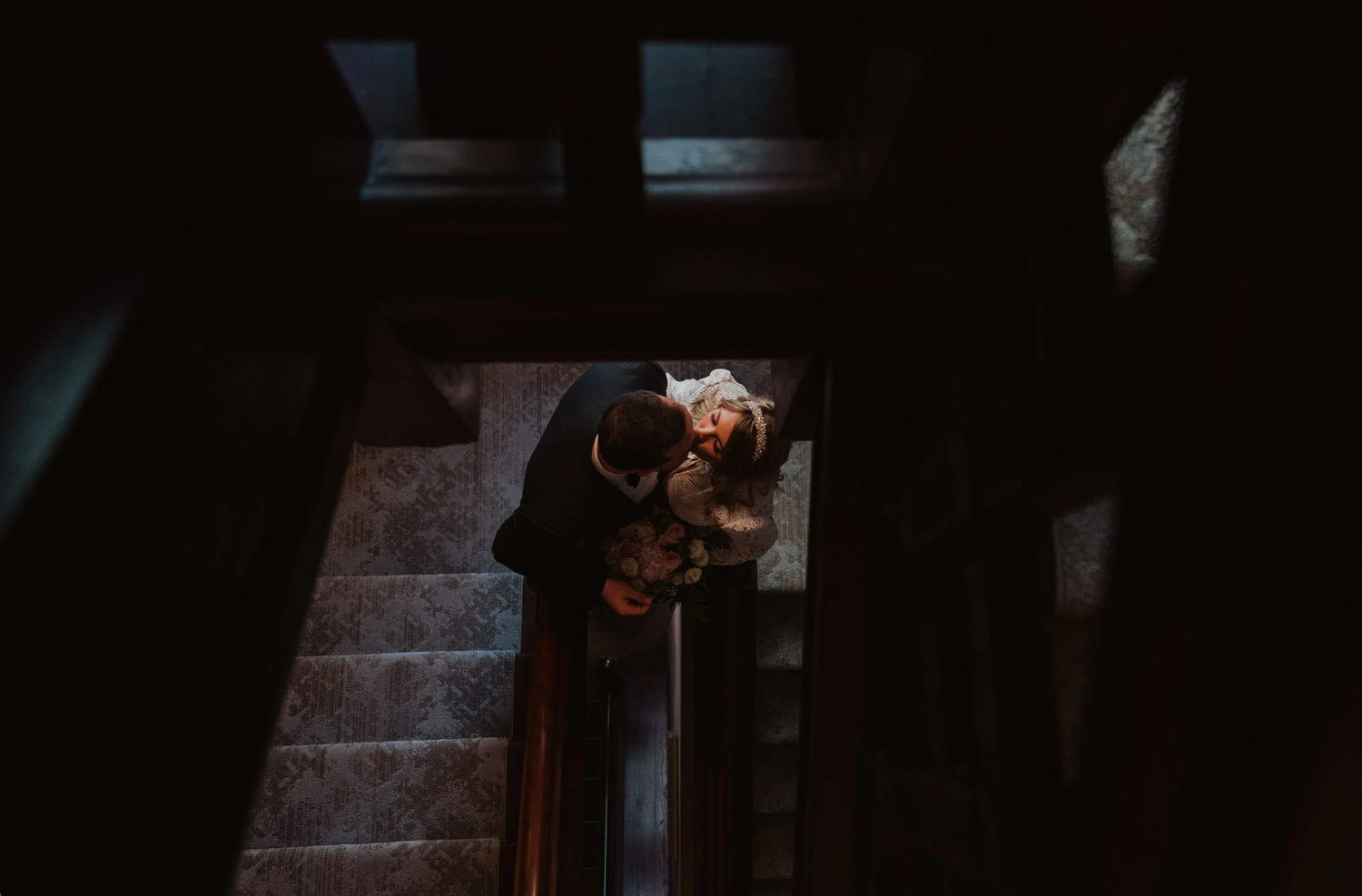 Two people embracing on a dimly lit staircase in a dark, moody interior setting.