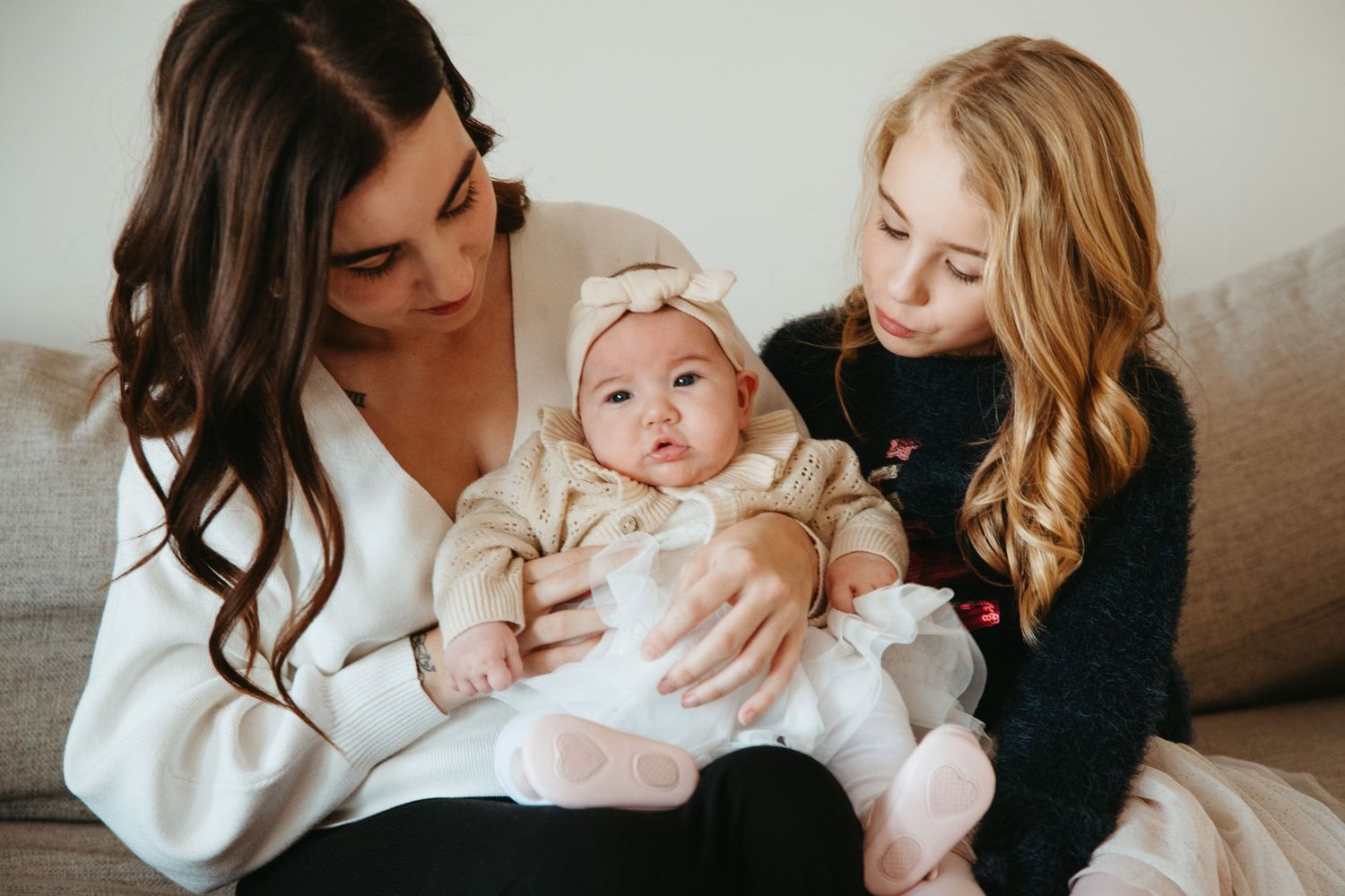 Two women sitting on a couch lovingly holding a cute baby girl wearing a bow headband.