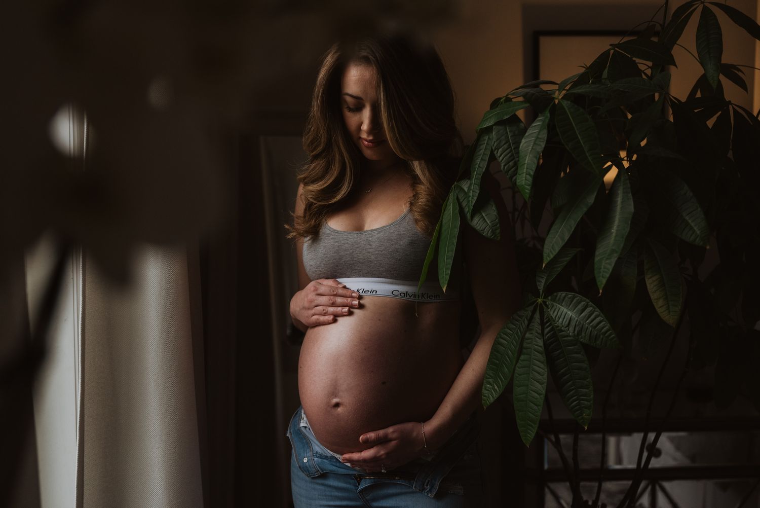 Pregnant woman in gray bra and jeans gently holding baby bump near green indoor plant.
