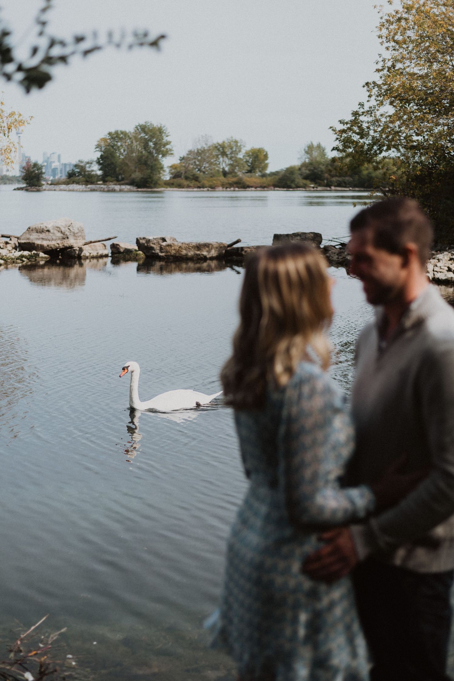 Couple standing by a serene lake with a white swan swimming in the background.