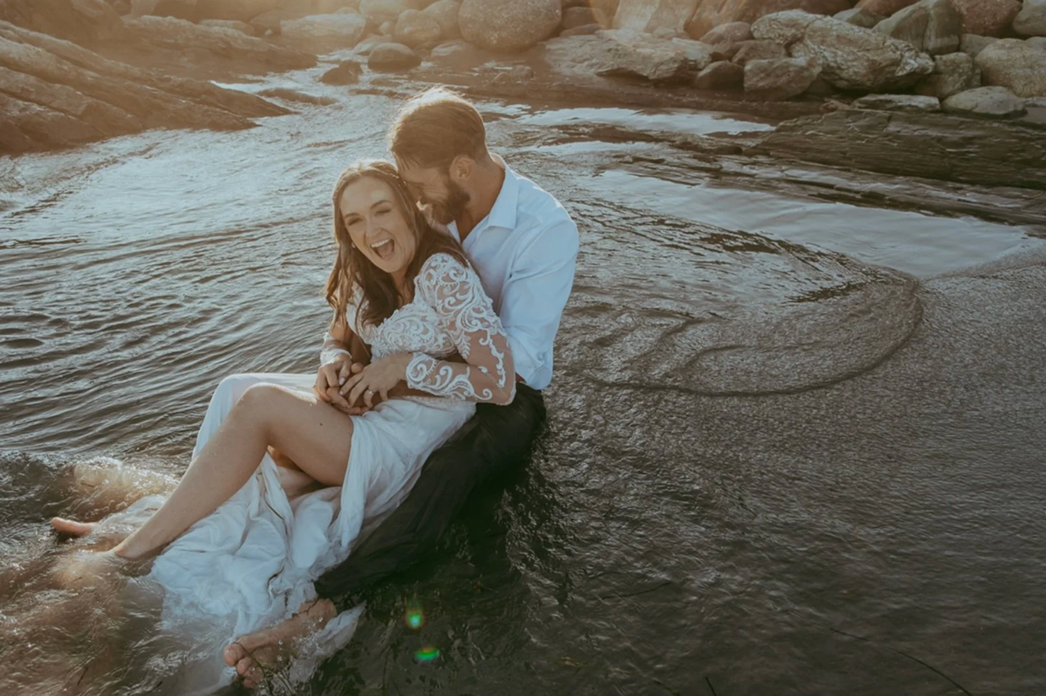 Romantic couple sitting together in shallow water near rocks, wearing white outfits, bathed in warm golden sunlight.