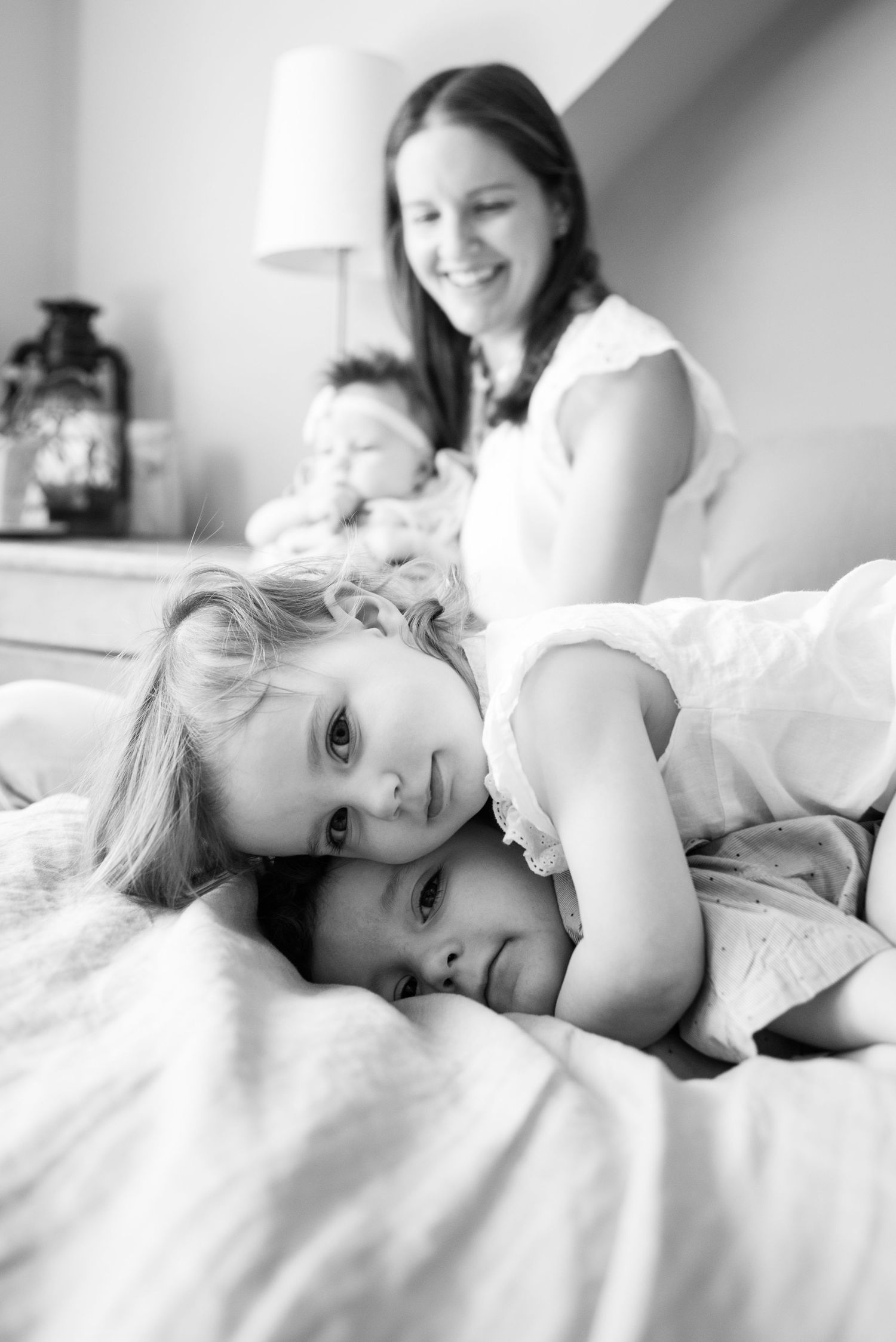 Black and white photo of two young girls lying on a bed while a smiling mother sits behind them.