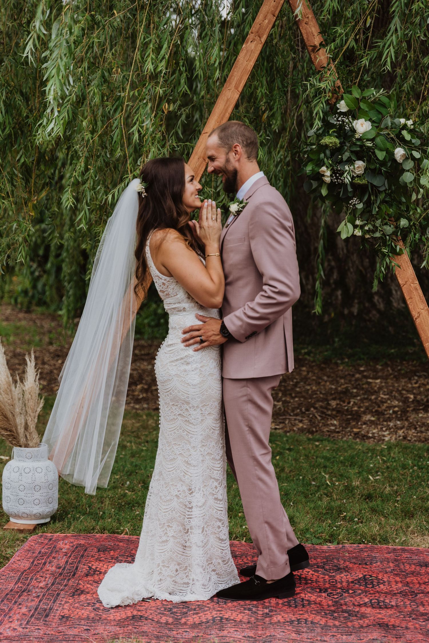 Bride and groom share a romantic moment under a triangular wooden arch with greenery at an outdoor wedding ceremony.