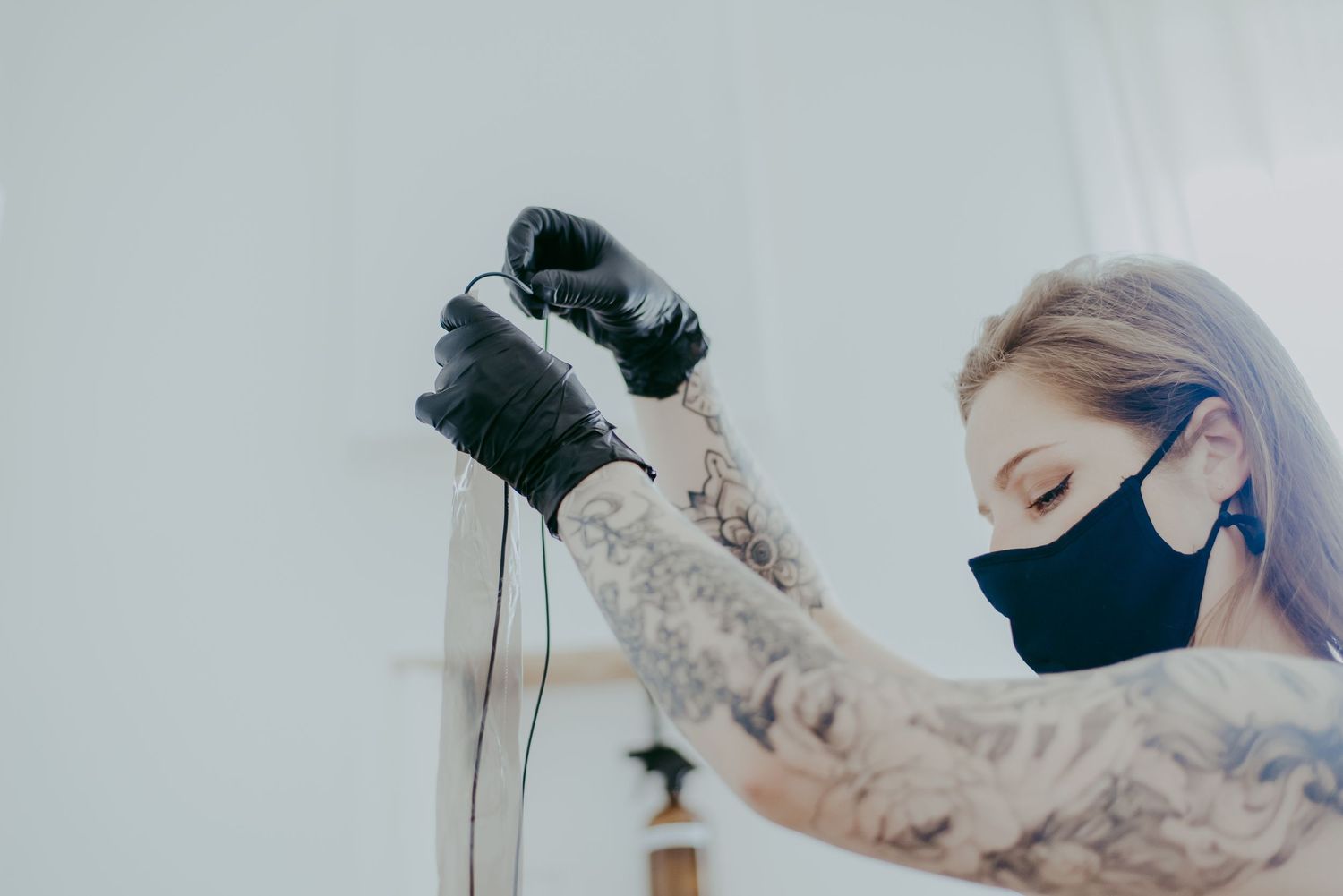 Tattooed woman wearing black gloves and face mask holds tattoo machine cord in a bright studio.