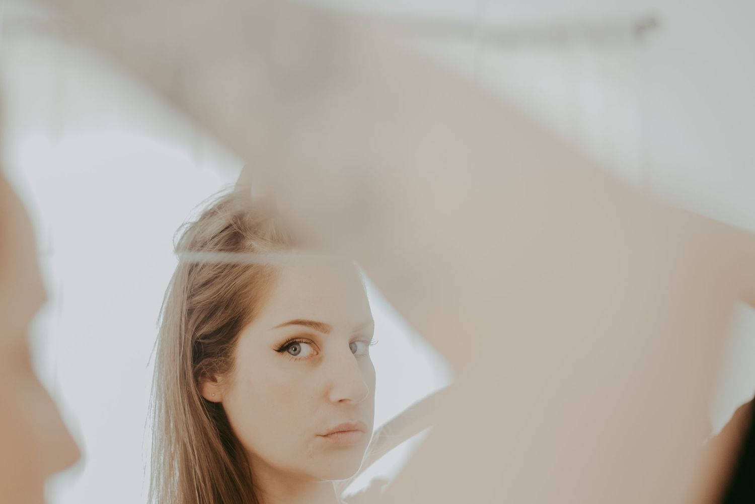 Young woman with long hair gazes into a mirror in a softly lit, ethereal white setting.