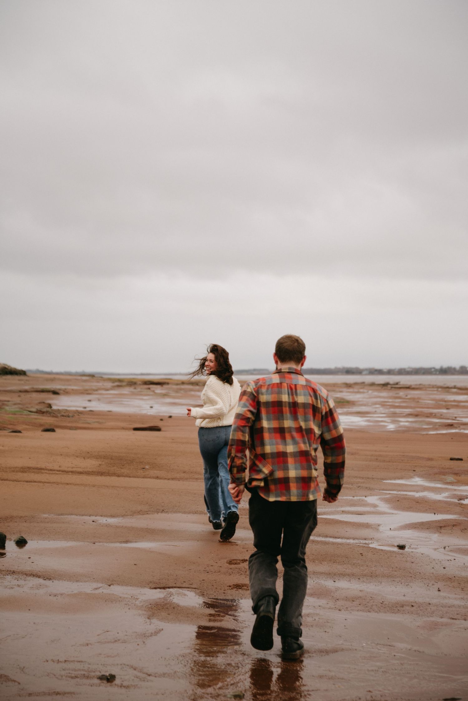 Couple walking hand-in-hand on a wet sandy beach on an overcast day, woman in white sweater and man in plaid shirt.