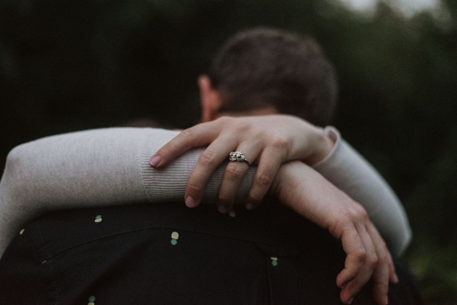 A couple embraces closely, showcasing an elegant engagement ring on the woman's hand resting on her partner's shoulder.