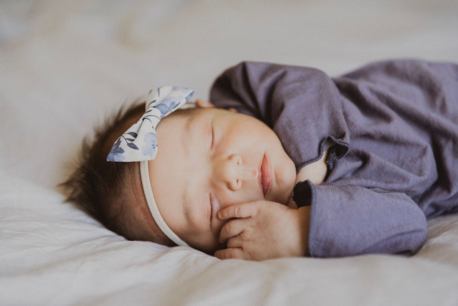 Newborn baby girl sleeping peacefully in purple outfit with white bow headband on white bedding.