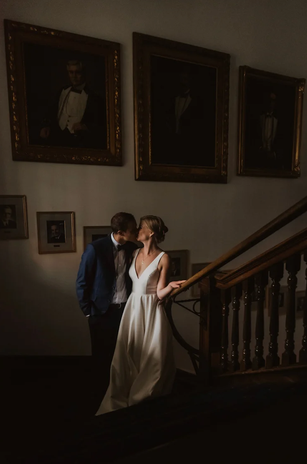 Bride and groom share a romantic moment on a grand staircase beneath golden-framed portraits in a historic venue.